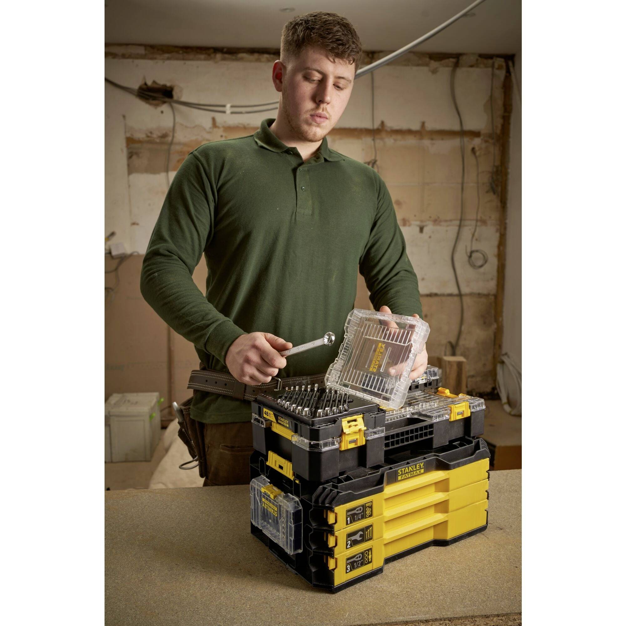 A man in a green shirt is standing in a workshop, holding a spanner while looking at a toolbox.