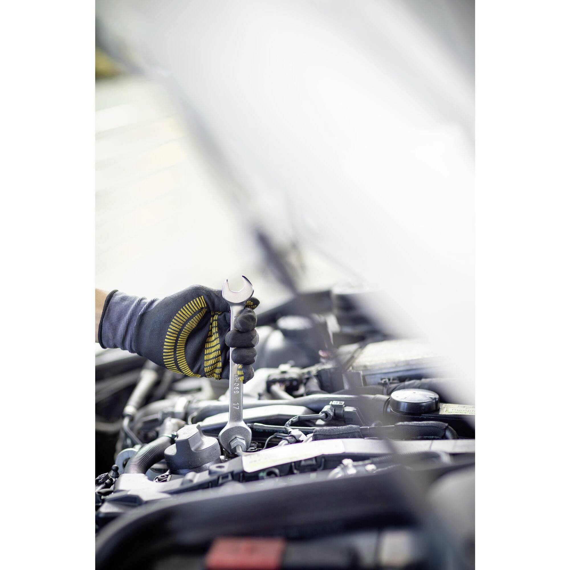 A mechanic's hand in a glove grips a spanner and works on a car's engine.