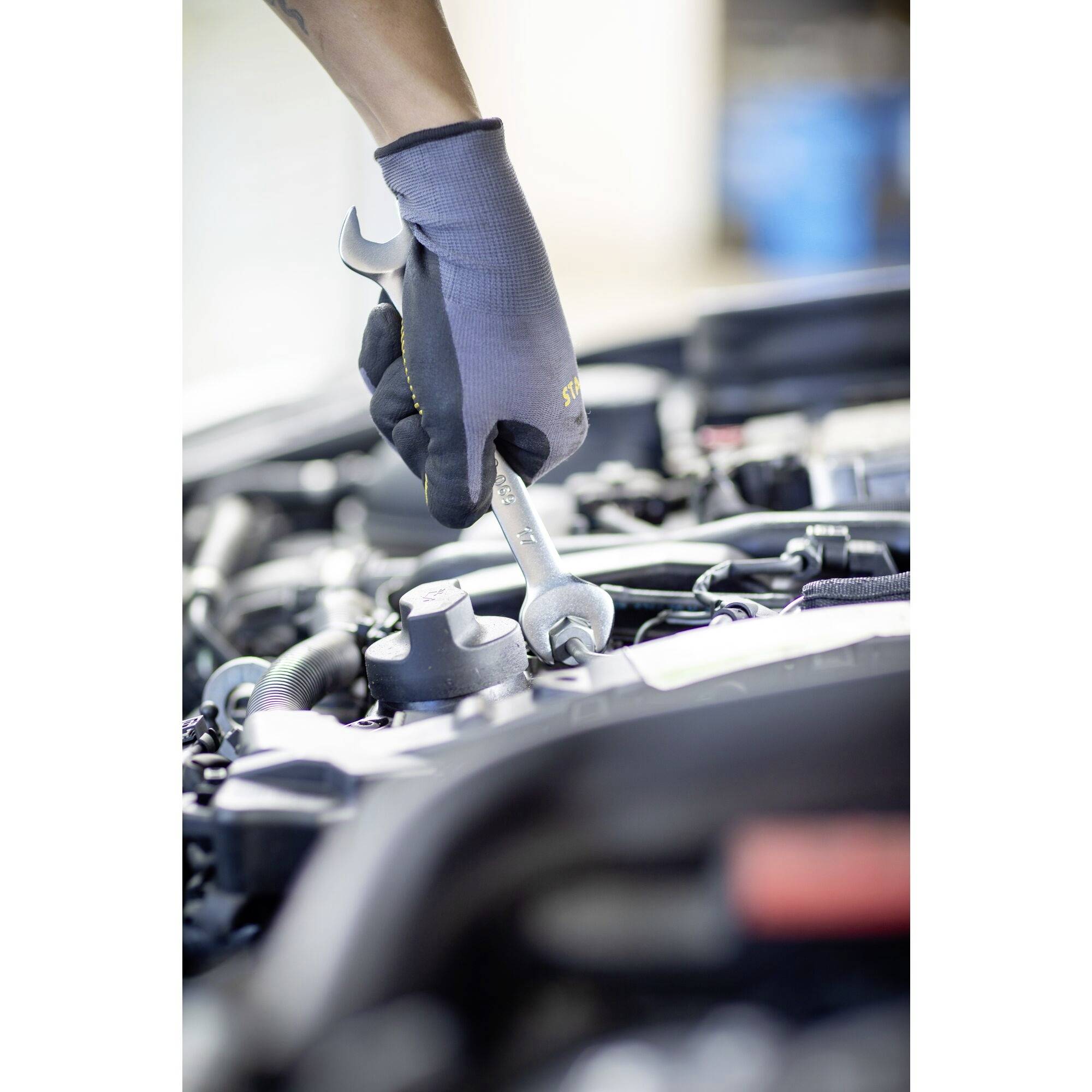 A person wearing gloves is working with a spanner on a car engine. A blurry workshop area is visible in the background.