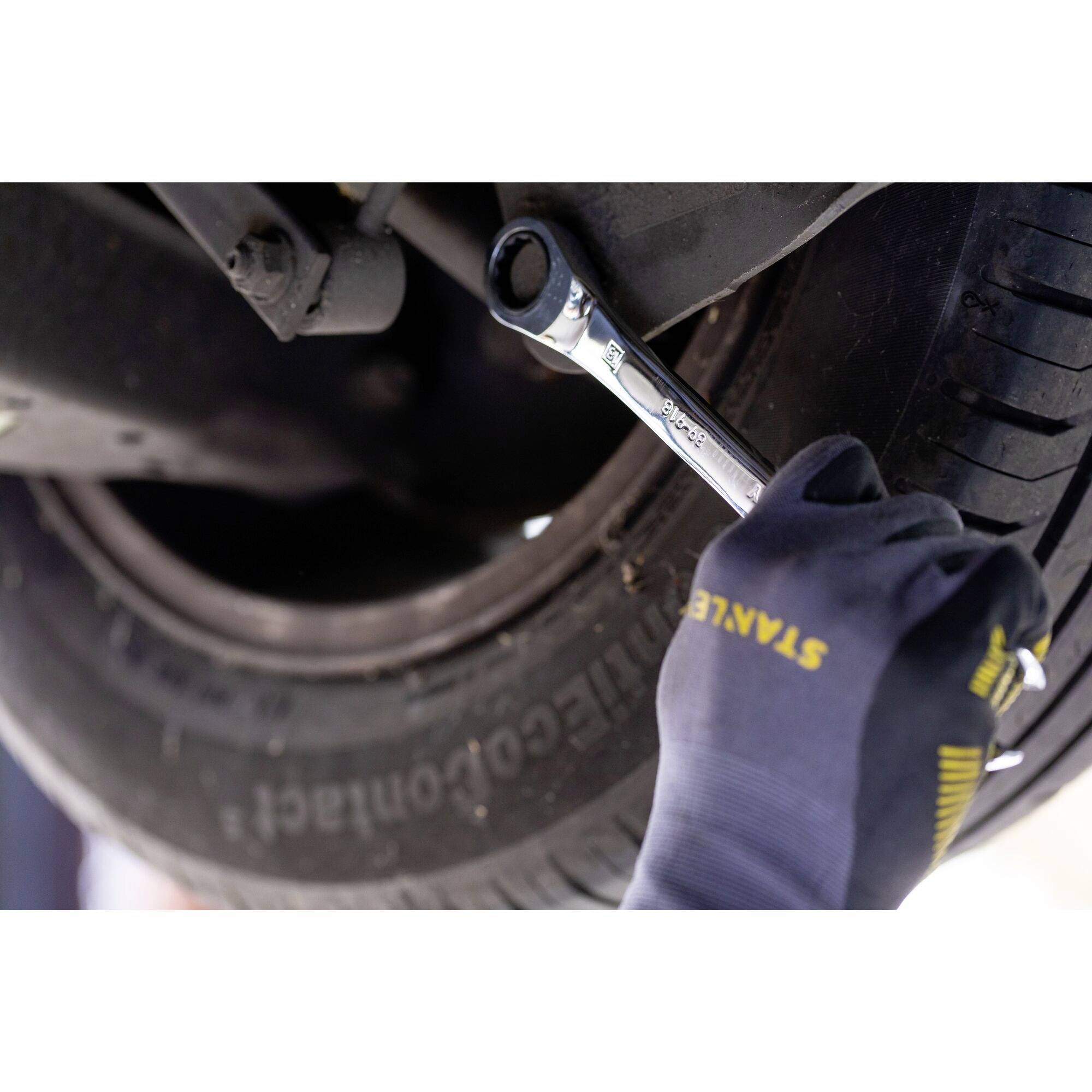 A gloved hand tightens a bolt on a vehicle wheel with a spanner. The focus is on the tool and wheel.