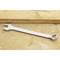 A silver ring spanner lies on a wooden table. The spanner has an open ring at one end and a closed ring at the other end.