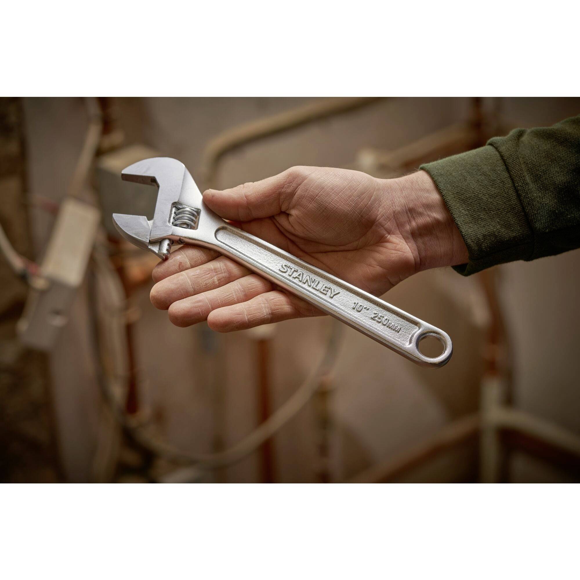 A hand holds an adjustable spanner in front of a blurred background with pipes.