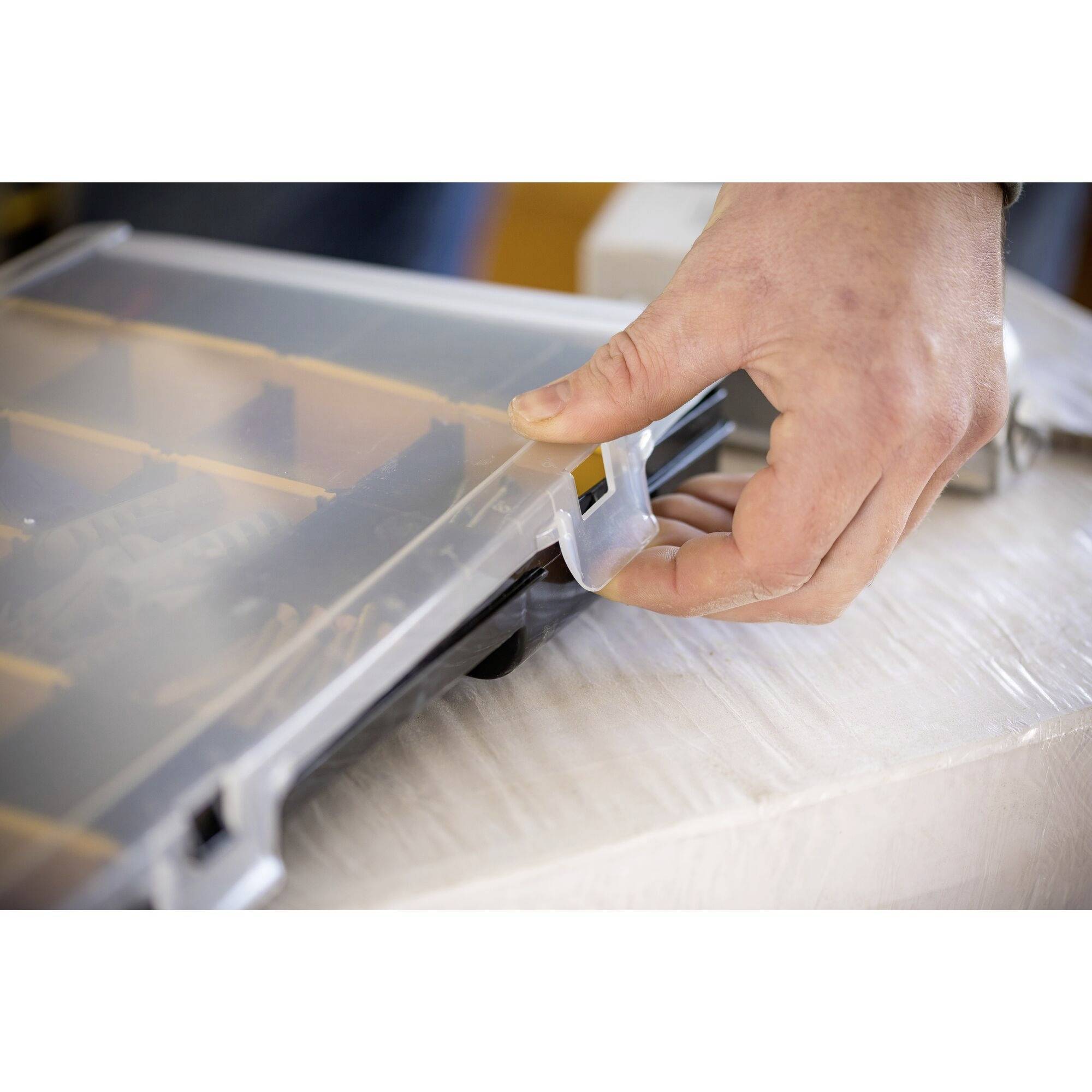 A hand is closing a transparent plastic box with multiple compartments, engaging the locking mechanism.