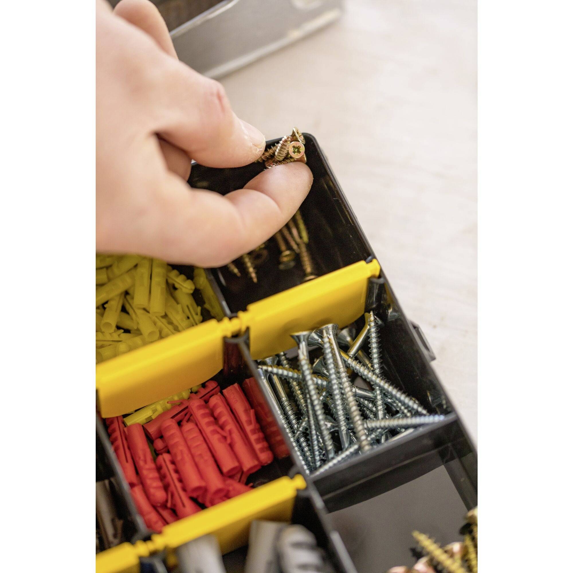 A hand takes a small screw from a sorting box filled with yellow wall plugs and silver screws.