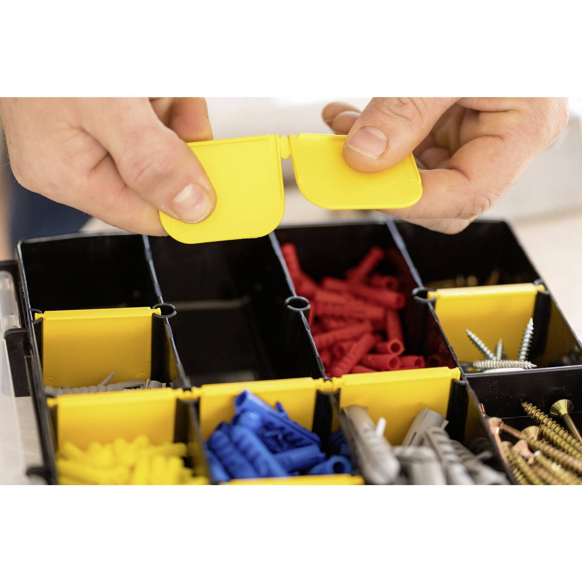 Close-up of hands opening the yellow dividers of a storage box. Colourful wall plugs and screws are neatly organised inside.