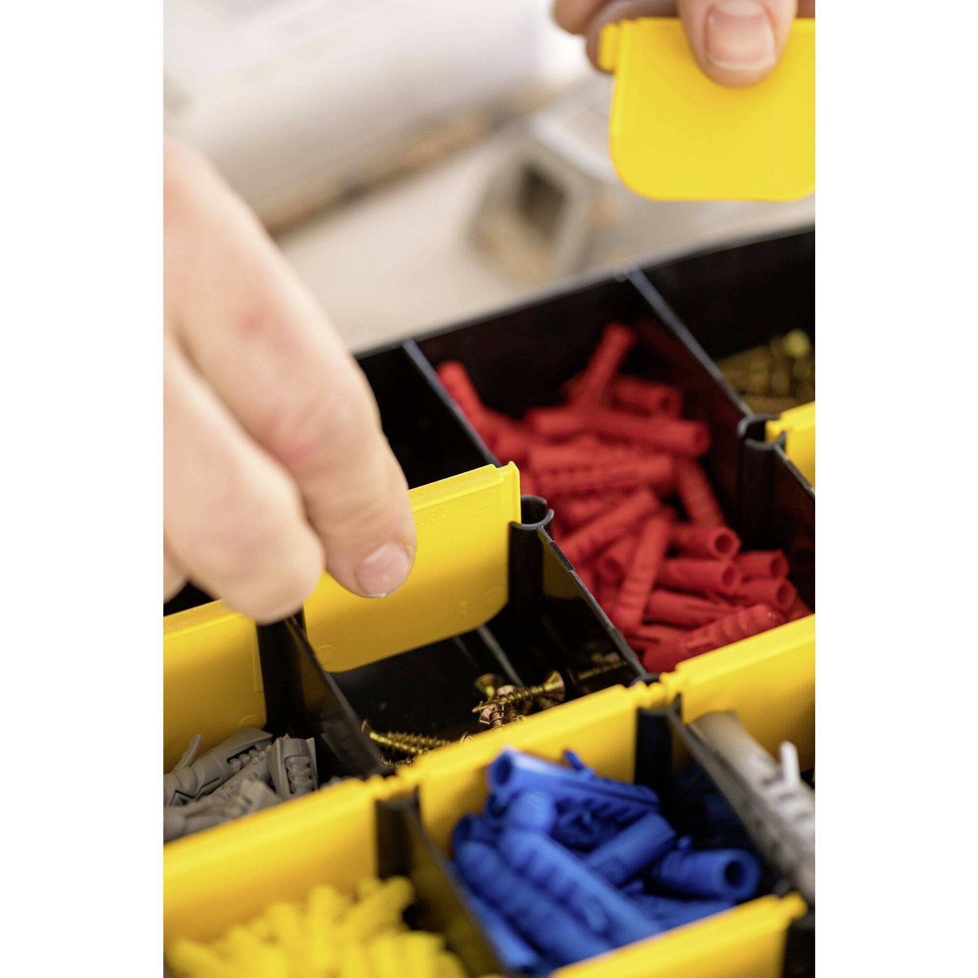 Close-up of hands organising a drawer with yellow, blue, and red wall plugs. One hand is holding a yellow divider card.