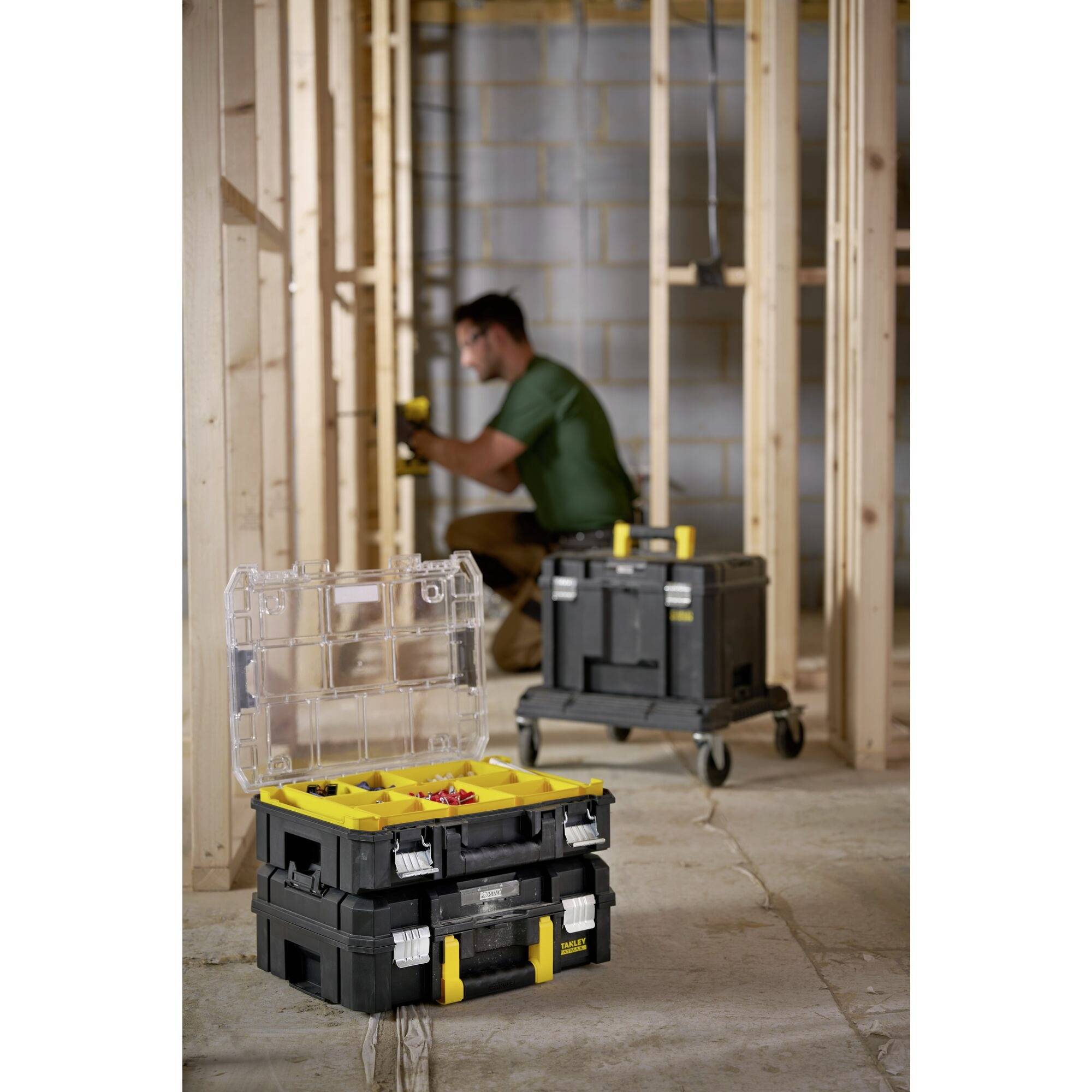 A tradesman on a building site, kneeling while working. In the foreground are two black tool boxes with yellow handles and open compartments.