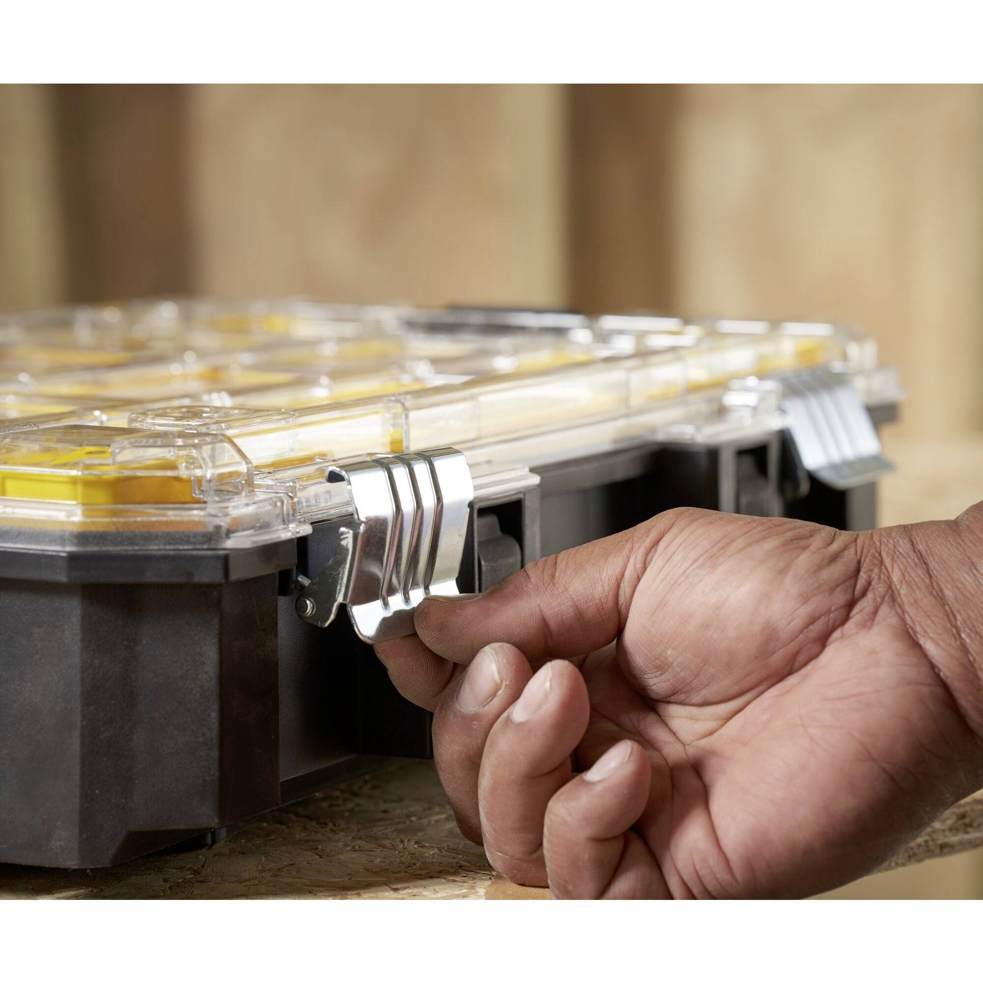 A hand opens the clasps of a transparent plastic toolbox with yellow compartments on a wooden table.