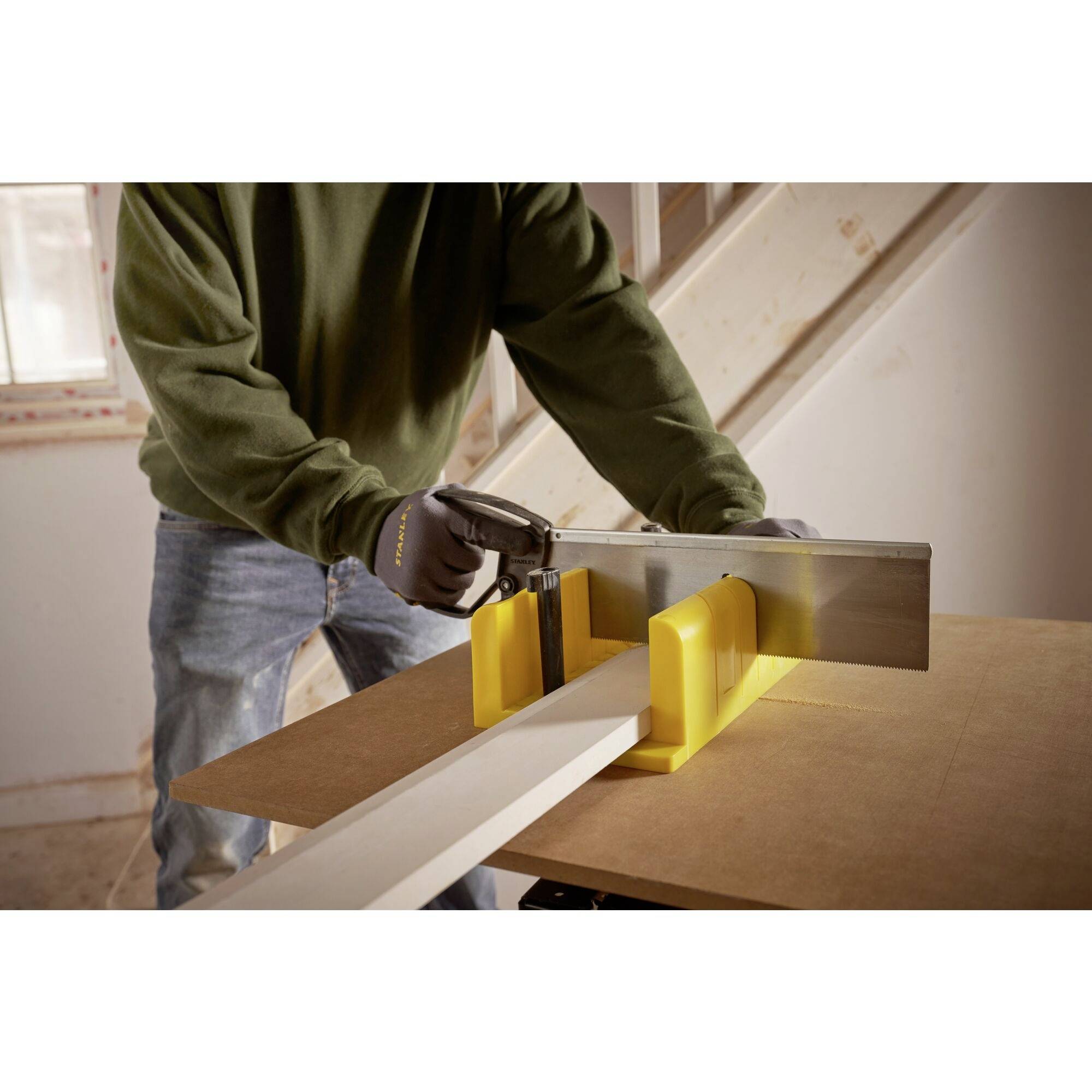 A person is sawing a wooden board in a workshop. They are wearing a green jumper and holding a saw guided by a yellow tool.