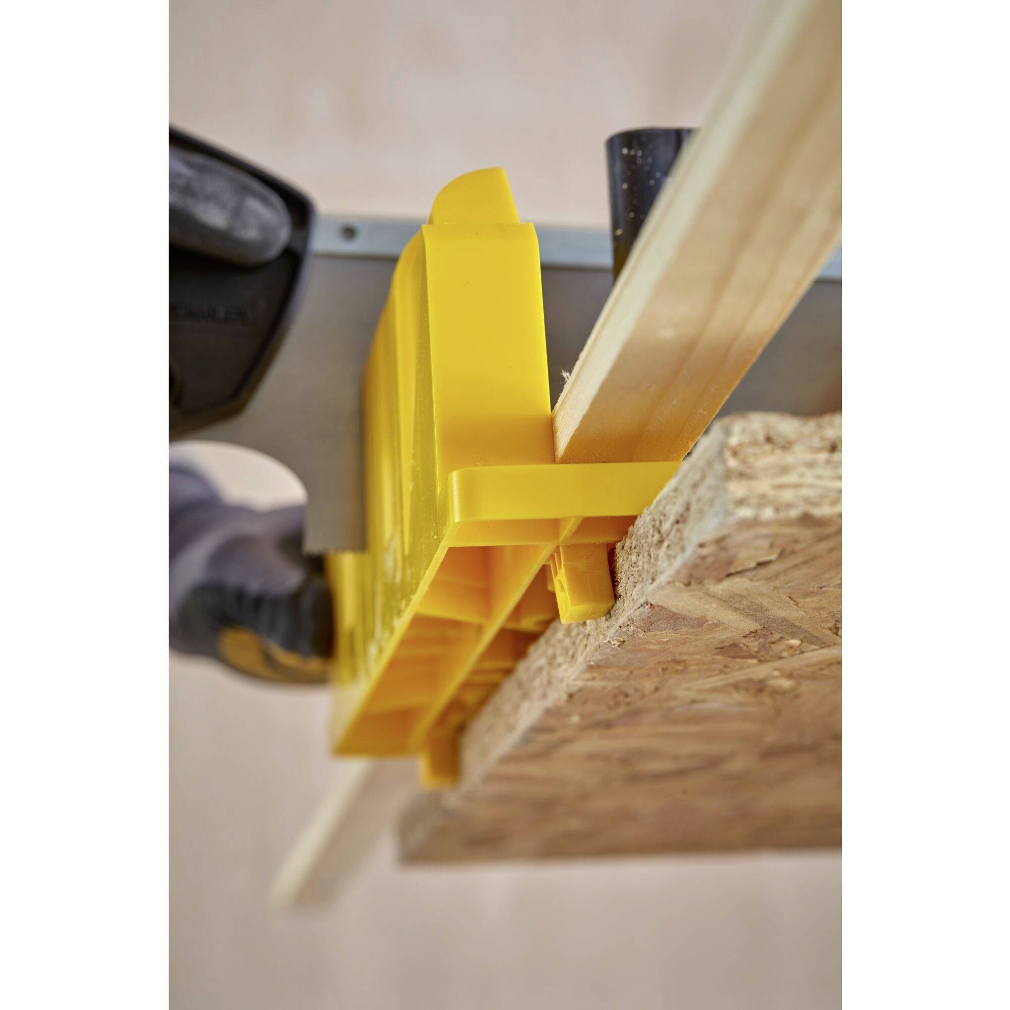 A yellow mitre box holds a piece of wood being cut with a saw. A centrally positioned hand stabilises the tool.
