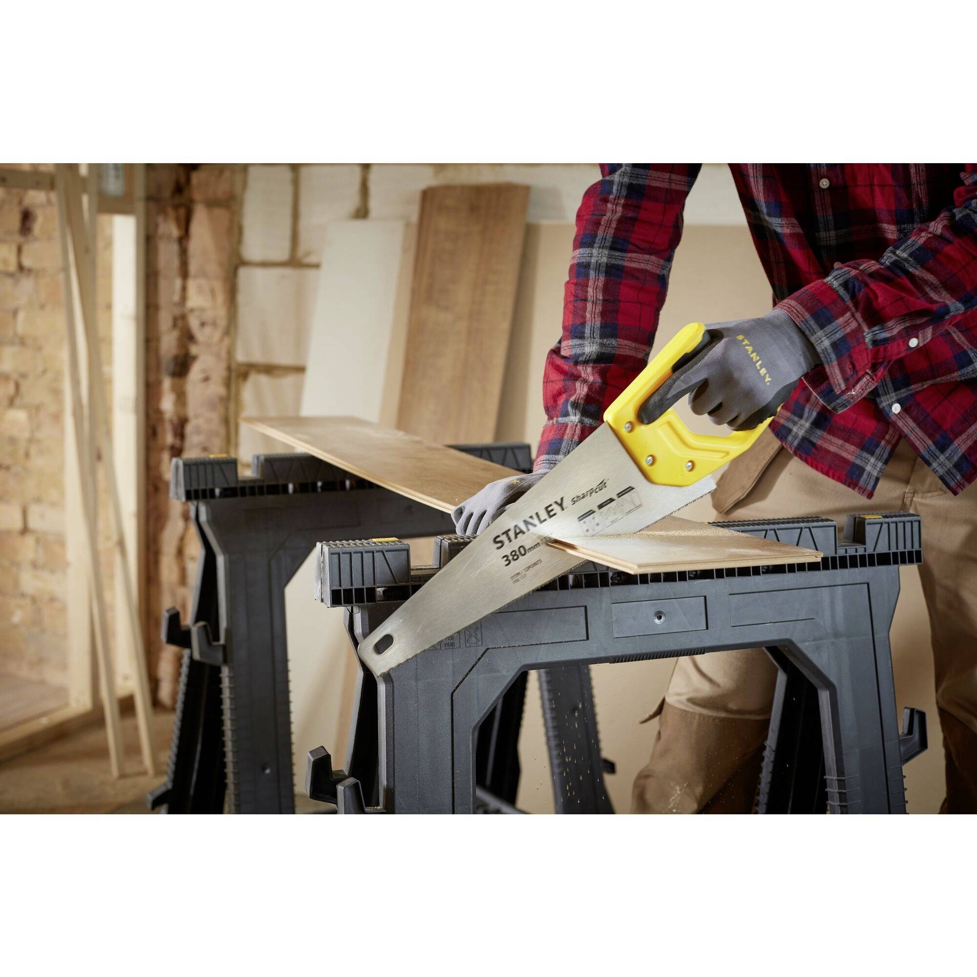 A person in workwear is sawing a wooden board on two sawhorses using a handsaw. Wooden planks are visible in the background.