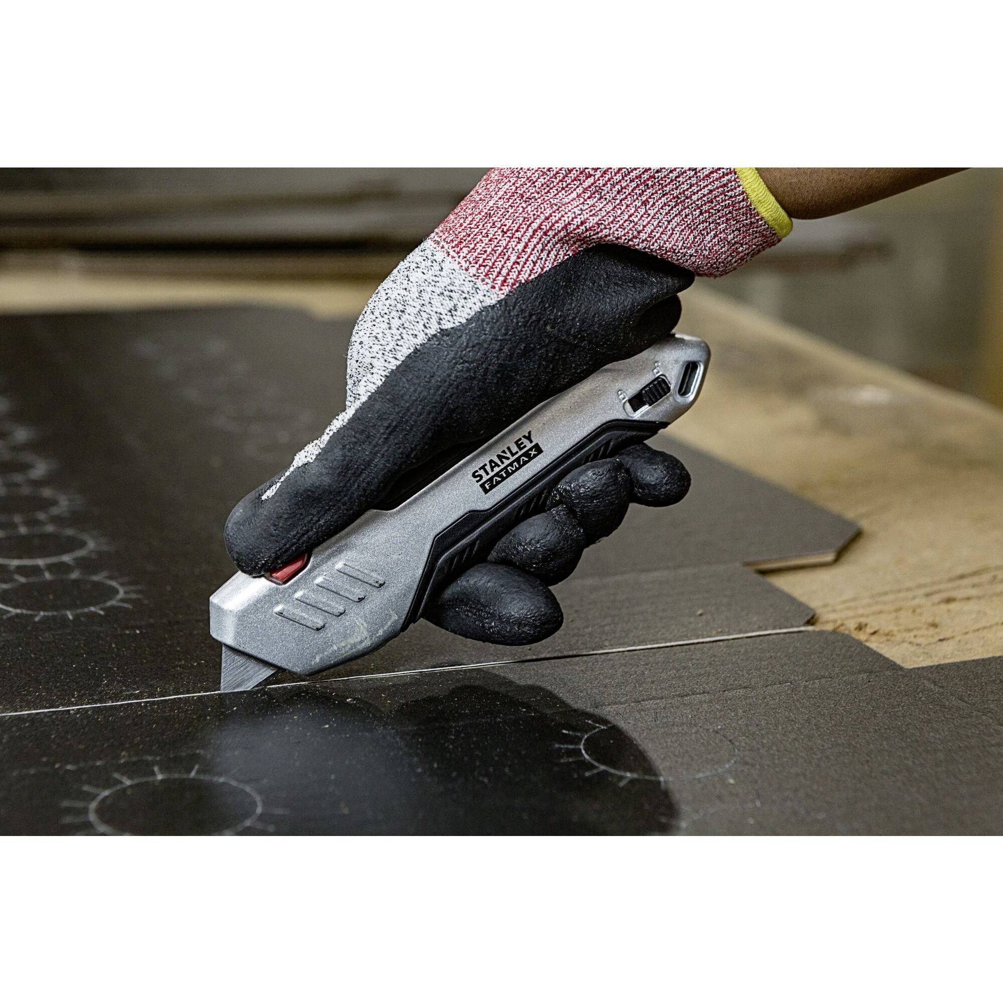 A hand wearing a work glove runs a Stanley craft knife across a dark surface to cut it.