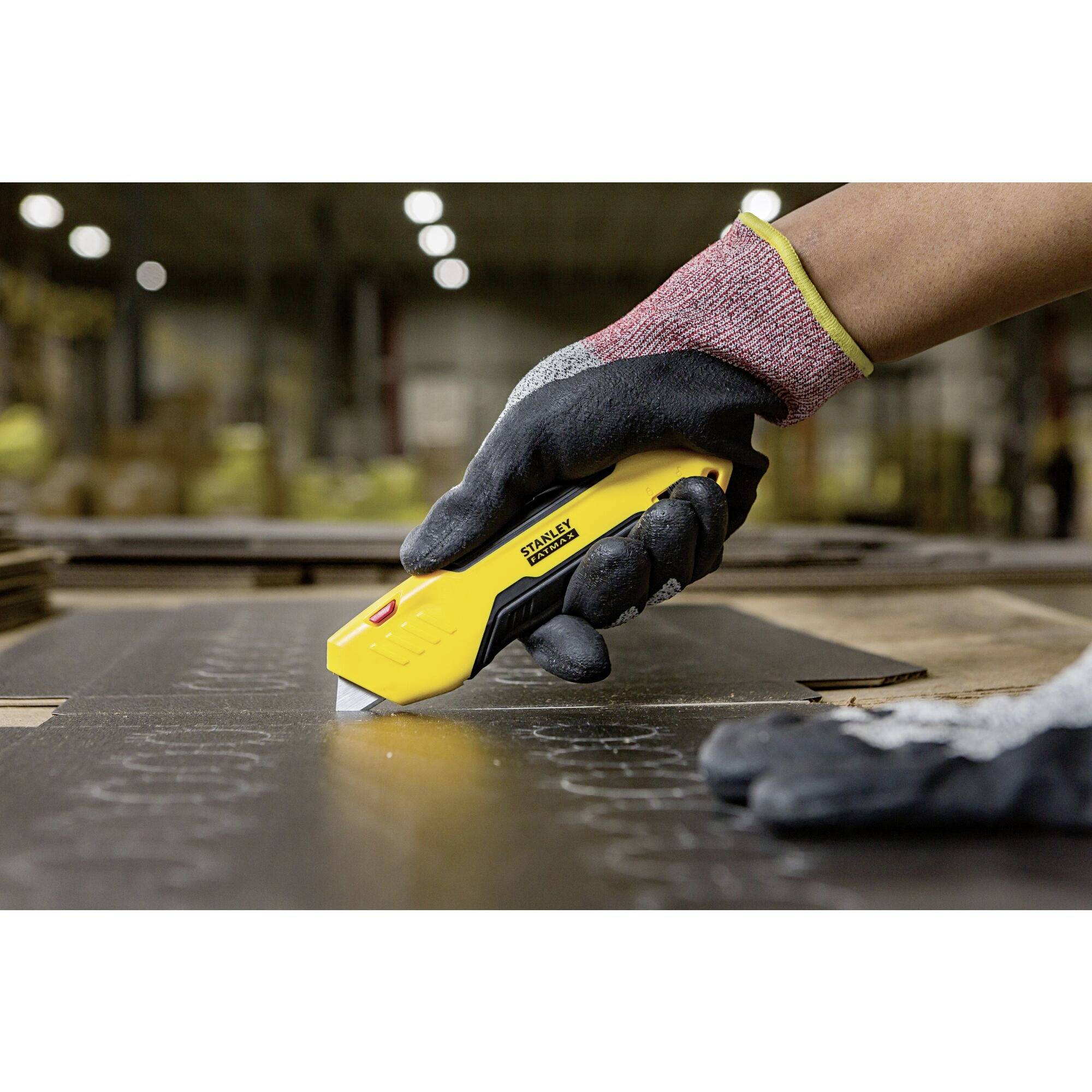 A gloved hand holds a yellow Stanley craft knife and cuts precisely through a black material in a warehouse setting.