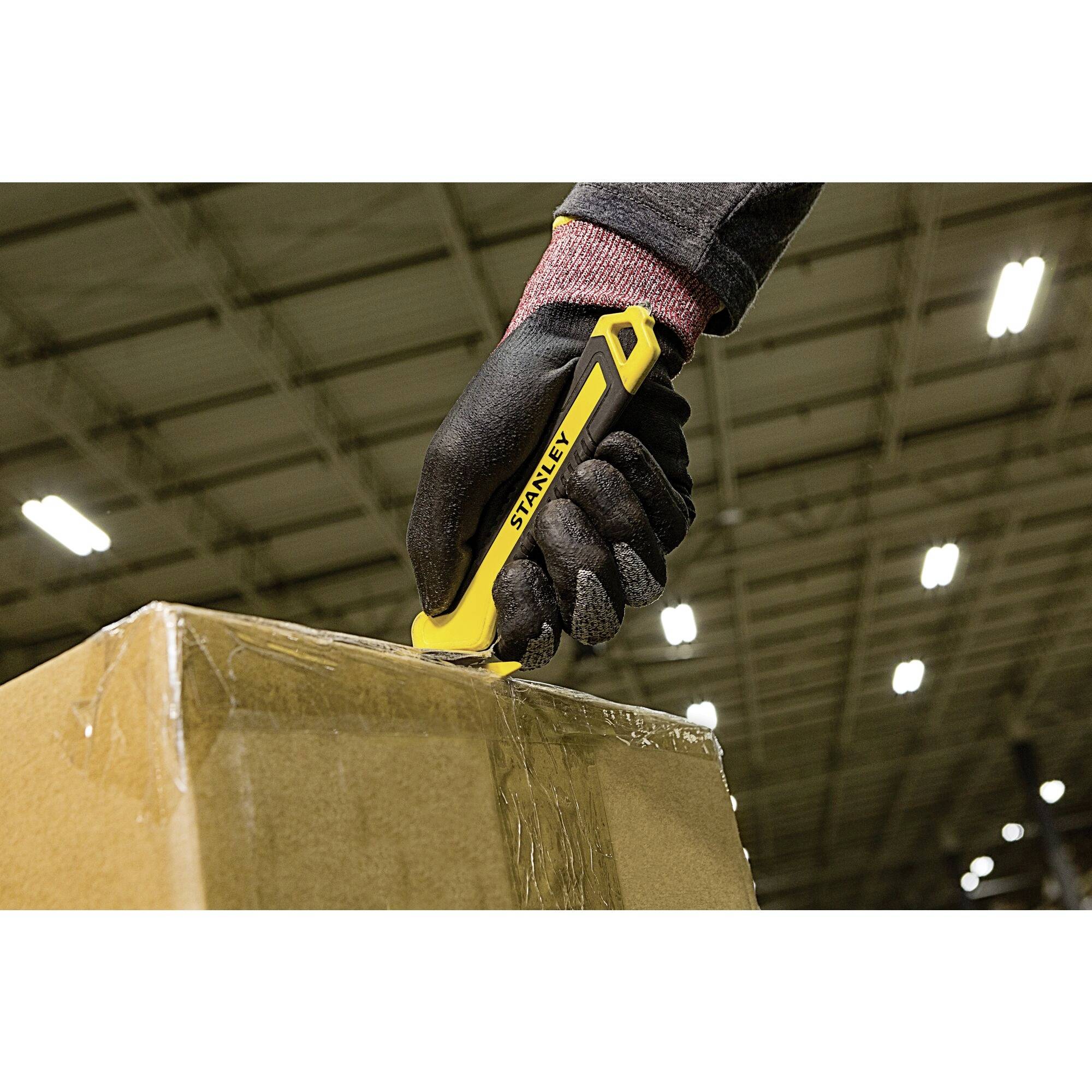 A person is cutting open a package with a yellow Stanley knife in a large warehouse. They are wearing black gloves.
