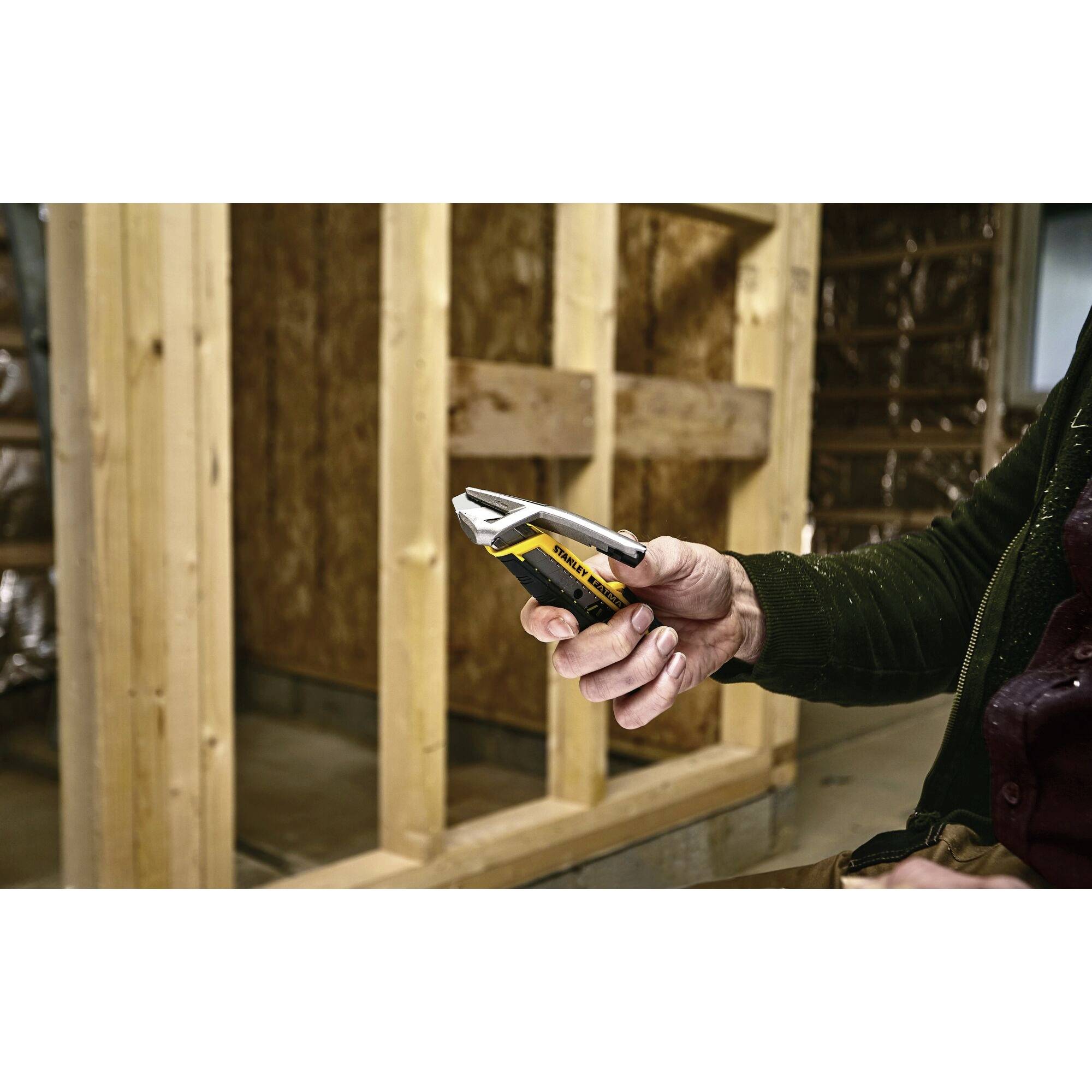 A person is holding a tape measure in front of a wooden wall in a building under construction. They are wearing a dark green jacket.