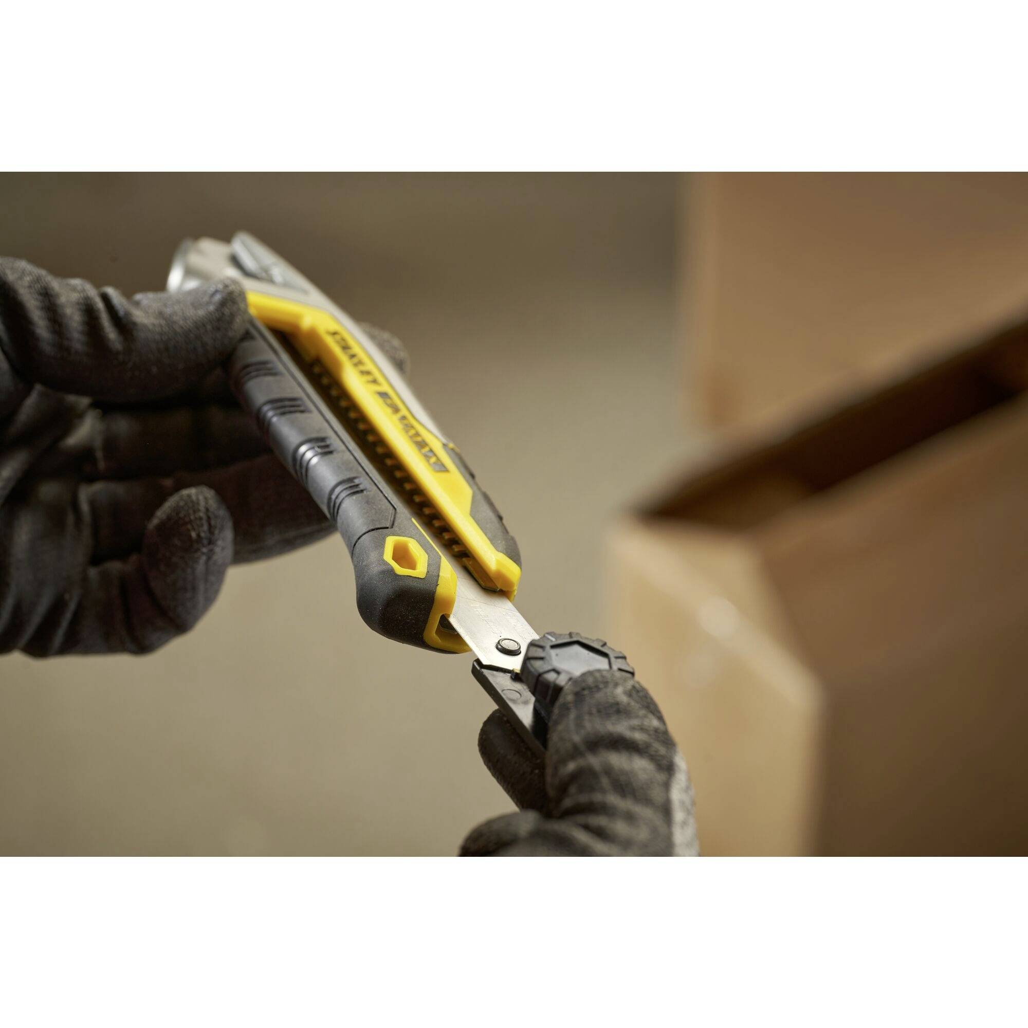 Two gloved hands are holding a yellow Stanley knife over a cardboard box, ready to cut. In the blurred background, an opened box is visible.