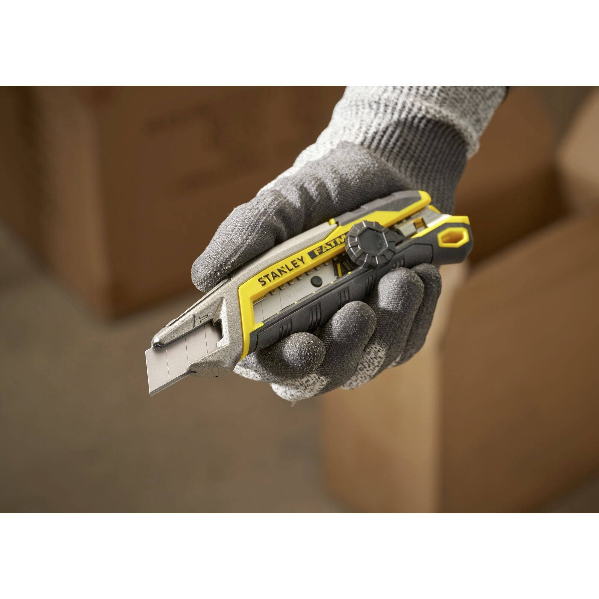 A gloved hand holds a yellow and grey utility knife with a retractable blade, surrounded by blurry cardboard boxes in the background.