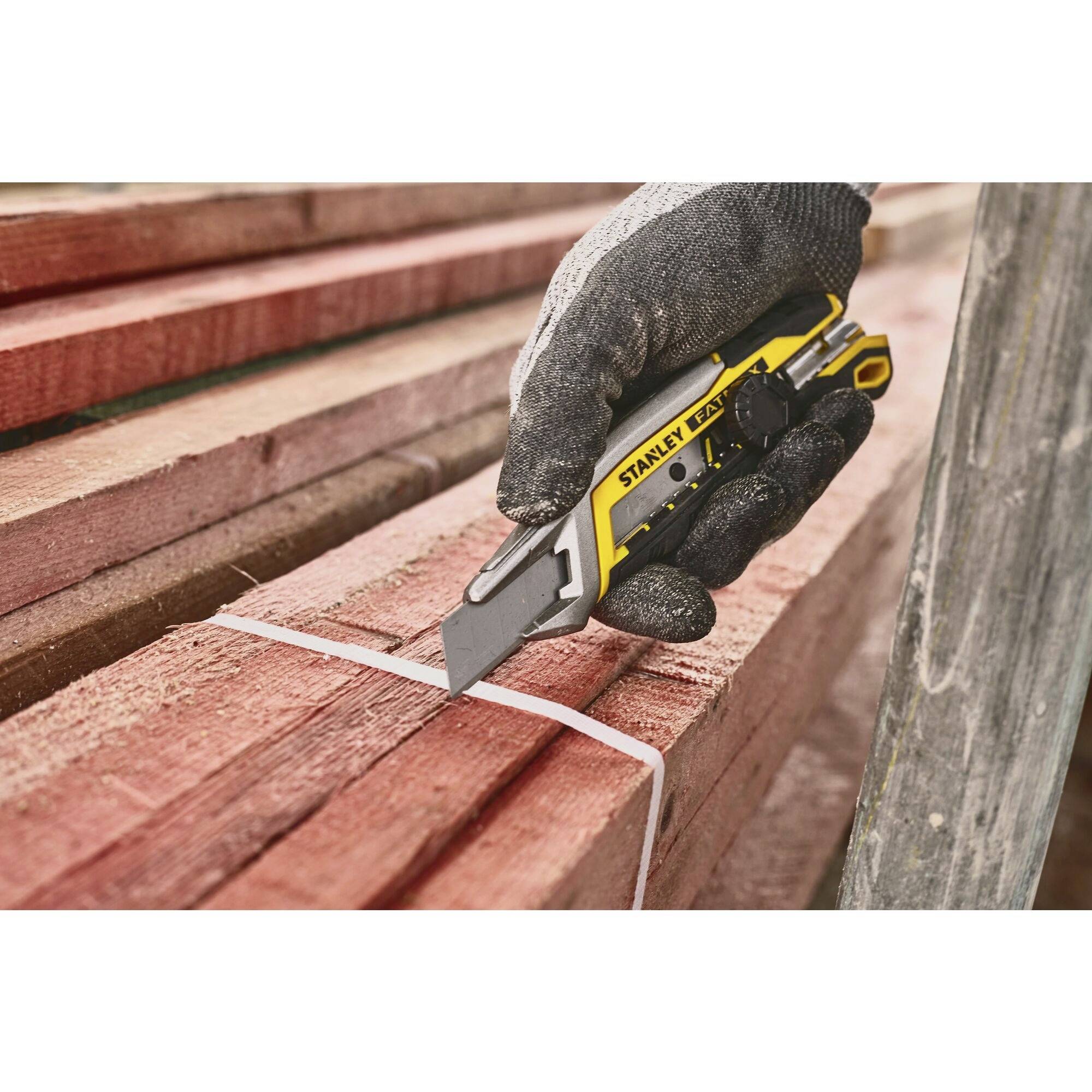 A gloved hand holds a utility knife to cut a plastic band around a stack of red wooden planks.