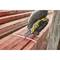 A gloved hand holds a utility knife to cut a plastic band around a stack of red wooden planks.