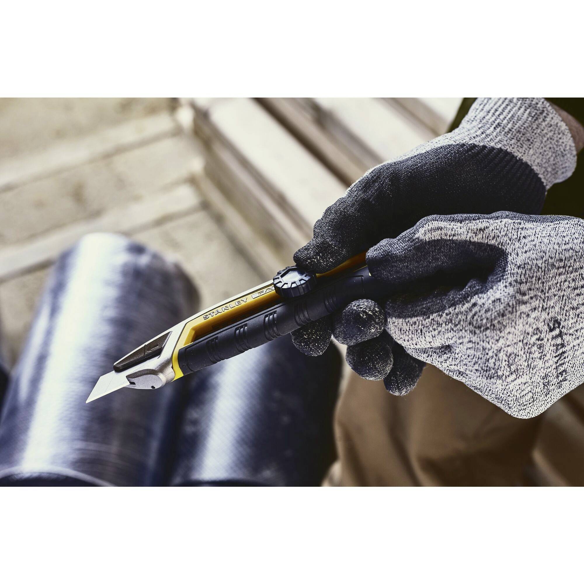 A person is holding a utility knife with a yellow blade, wearing grey work gloves. Black foil rolls are visible in the background.
