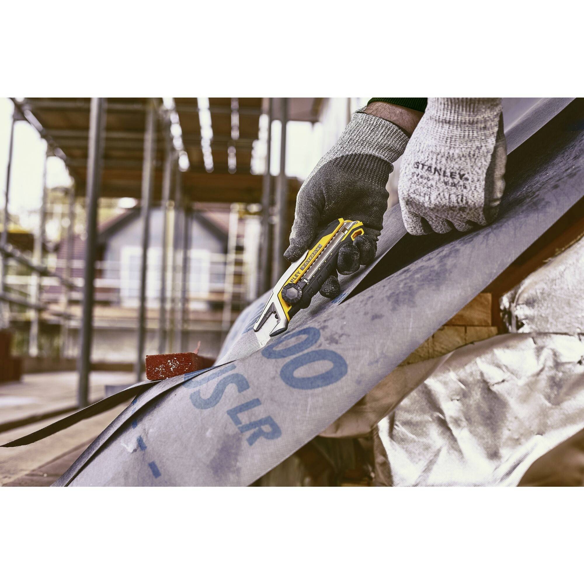 A person wearing work gloves is cutting a large, curved metal plate with a yellow utility knife on a construction site.