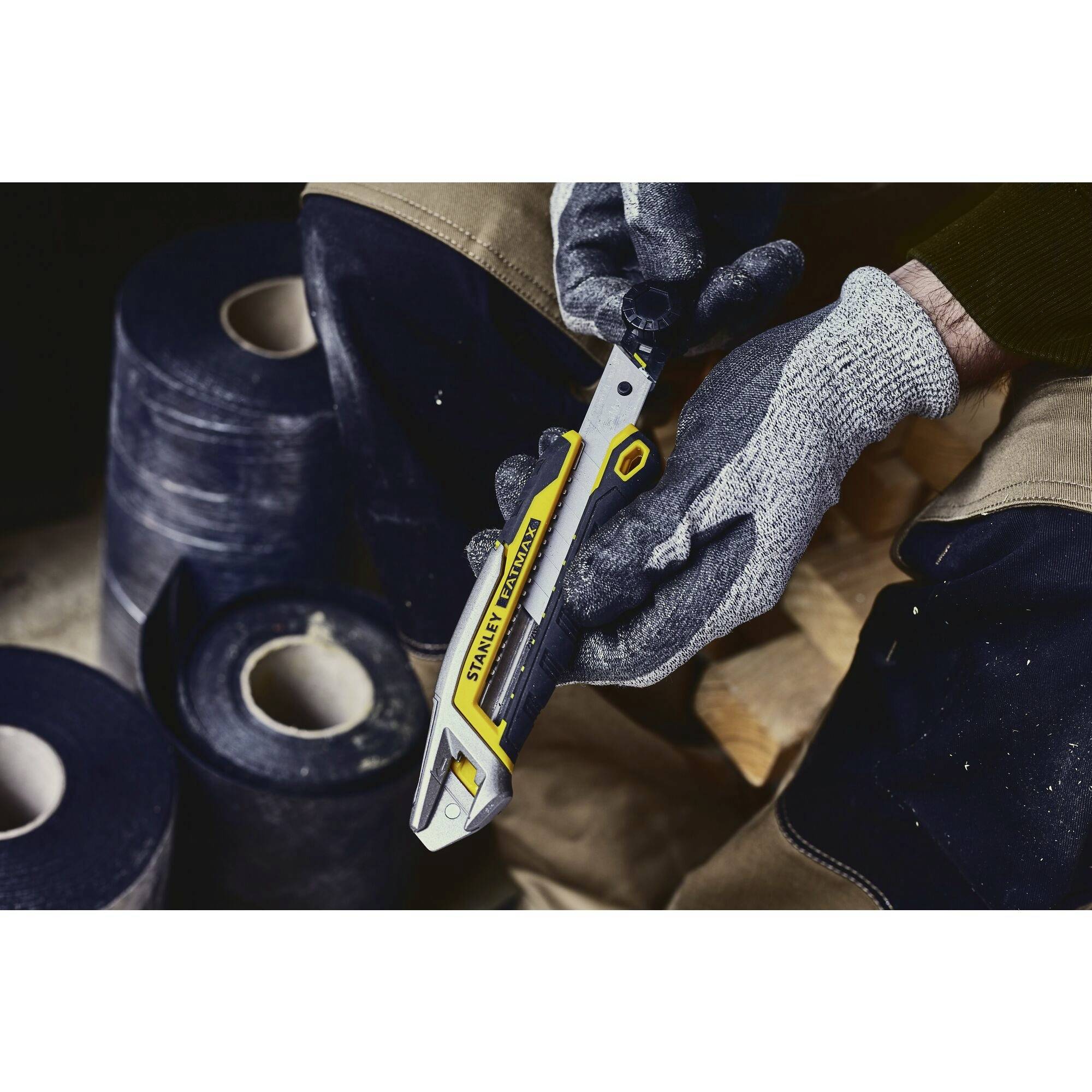 A person is wearing grey gloves and holding a yellow multi-tool. The background shows several black rolls.