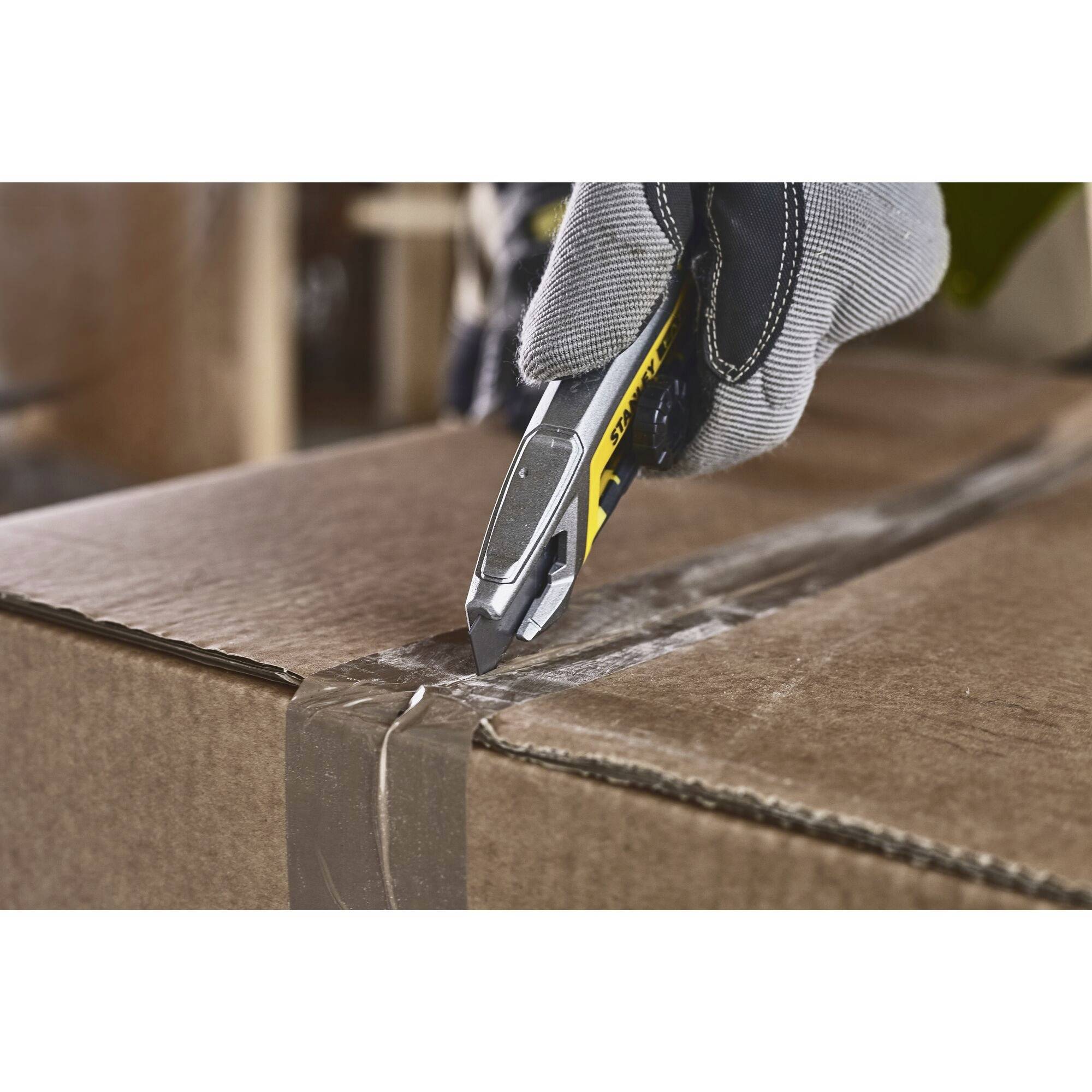 A hand wearing a work glove cuts adhesive tape on cardboard using a craft knife.
