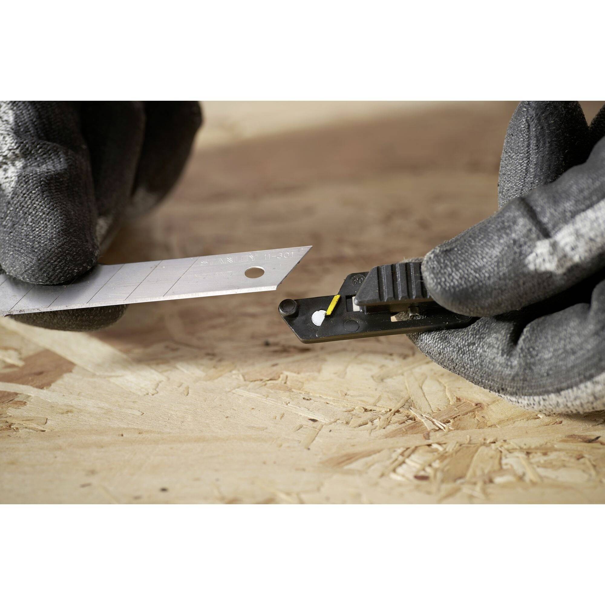 A person is holding a carpet knife and a spare blade with gloved hands over a wooden surface.