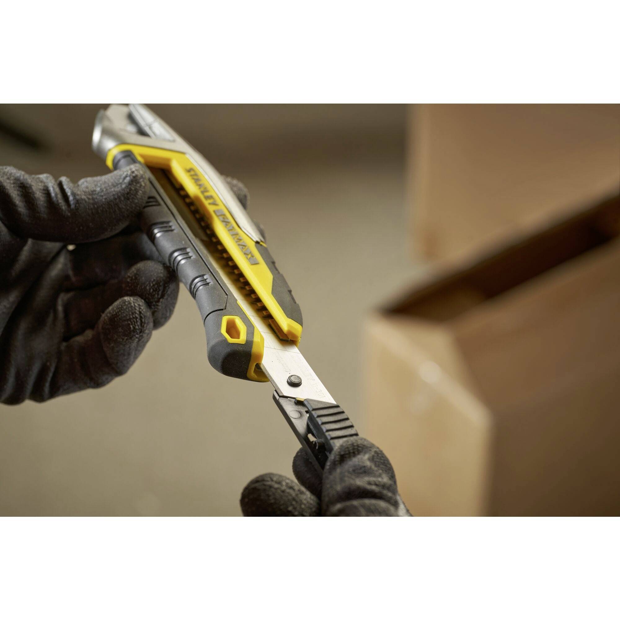 A person wearing black gloves is holding a yellow carpet knife. A cardboard box can be seen in the background.