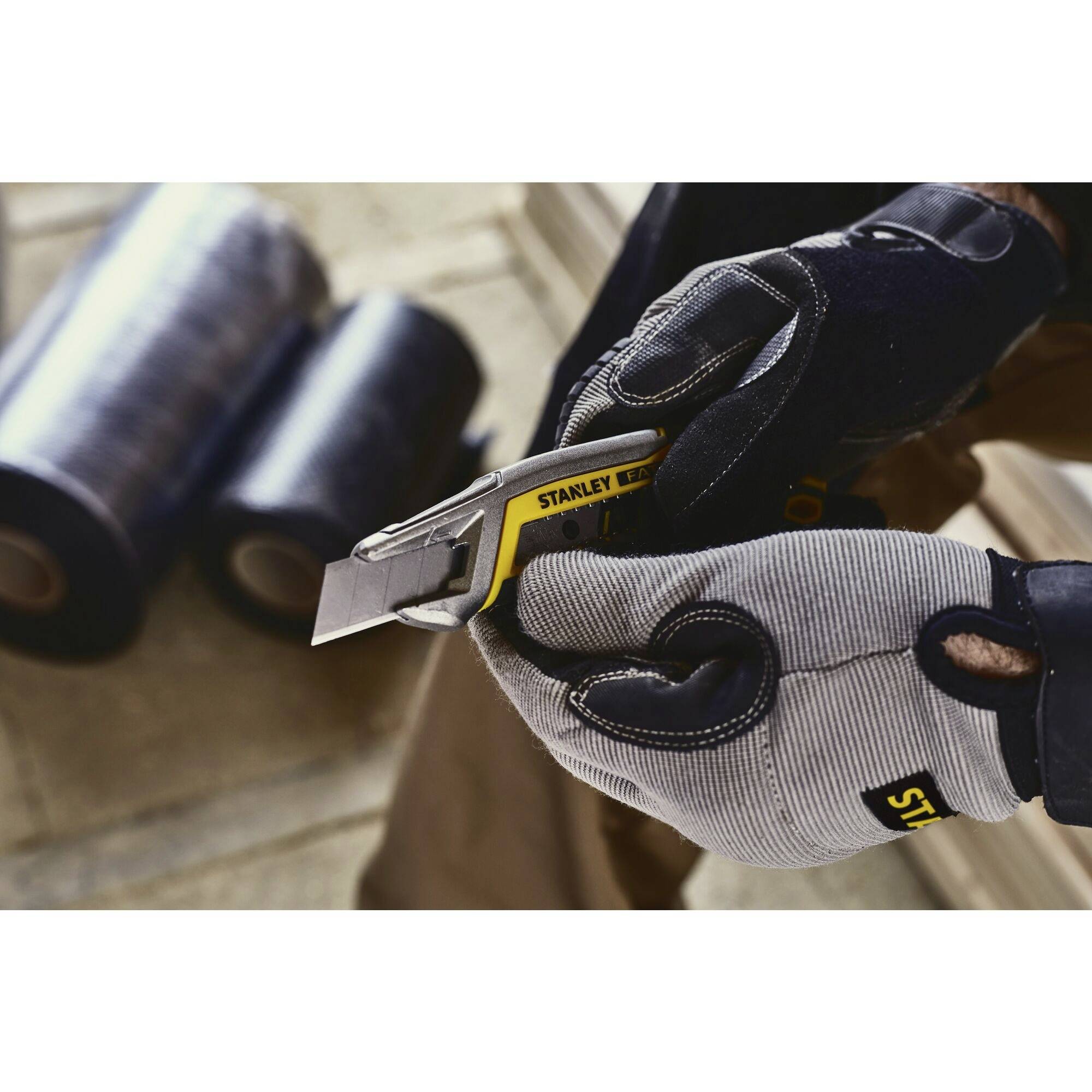 A person wearing gloves holds a safety knife in front of two rolls of packaging film. Workplace scenario; focus on safety.