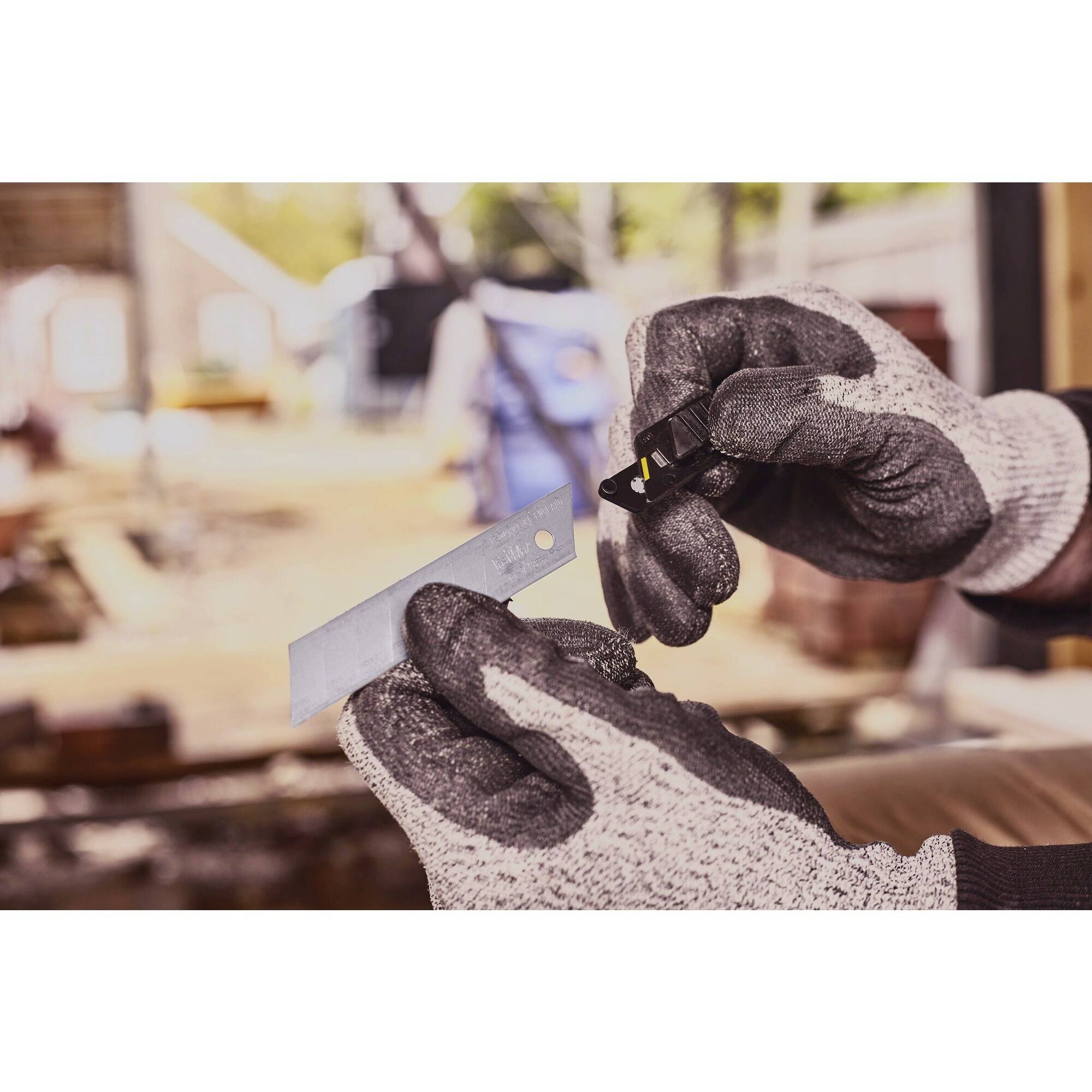 A hand wearing gloves grips a craft knife with a blade inserted; blurred construction site background.