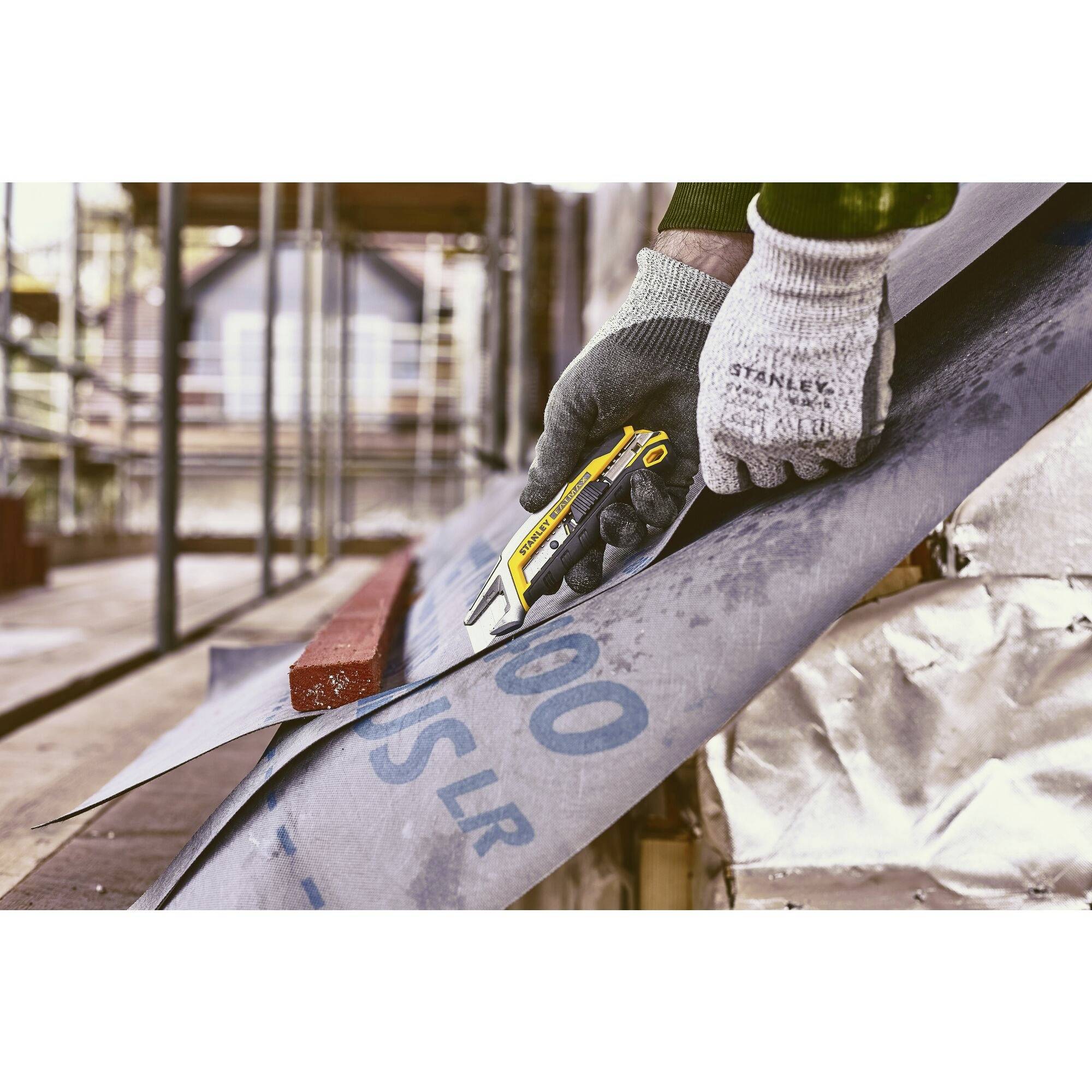 A person wearing gloves is cutting a large sheet of plastic with a craft knife in a construction site environment. Scaffolding can be seen in the background.