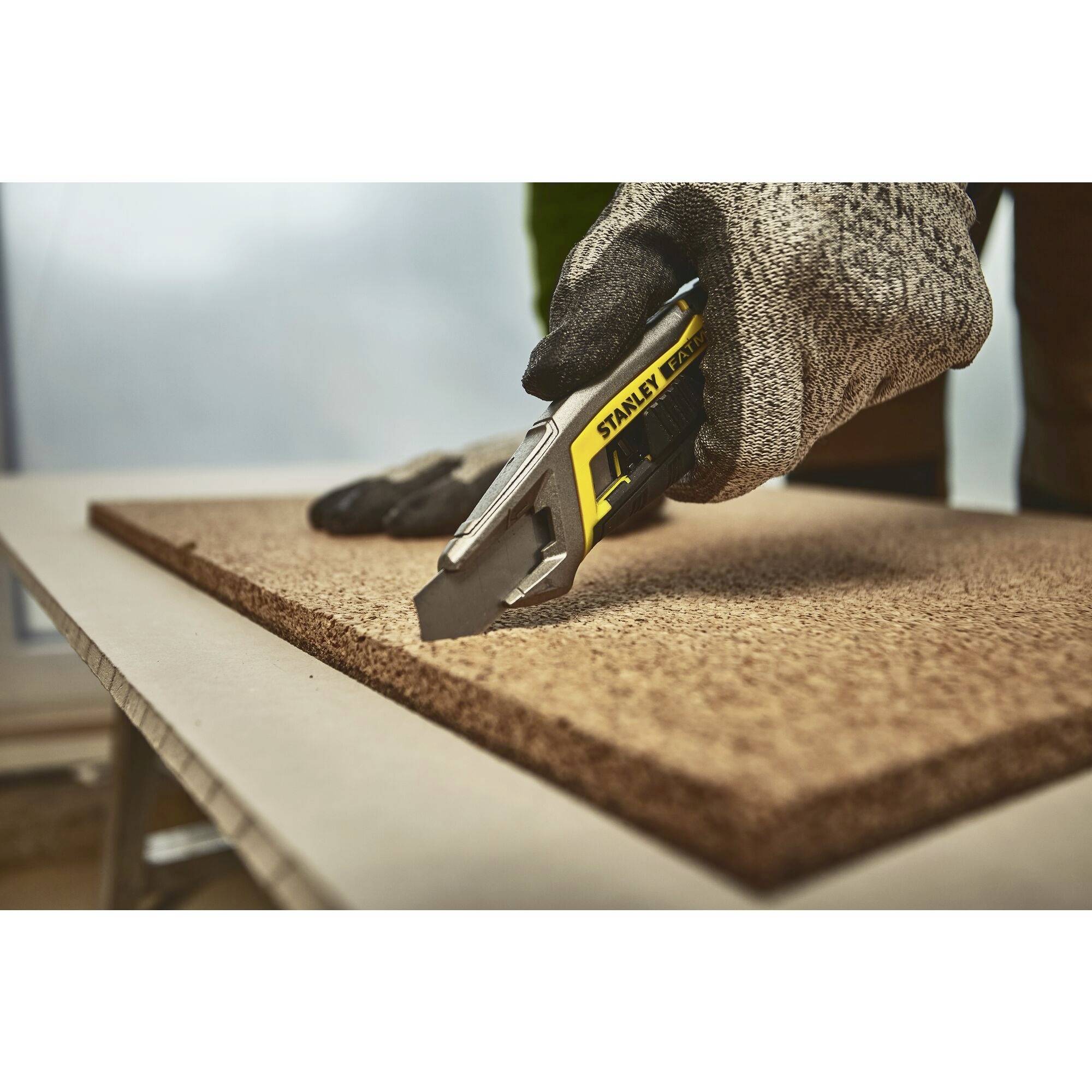 A person is cutting a piece of cork board with a craft knife on a table. Their hands are protected with work gloves.