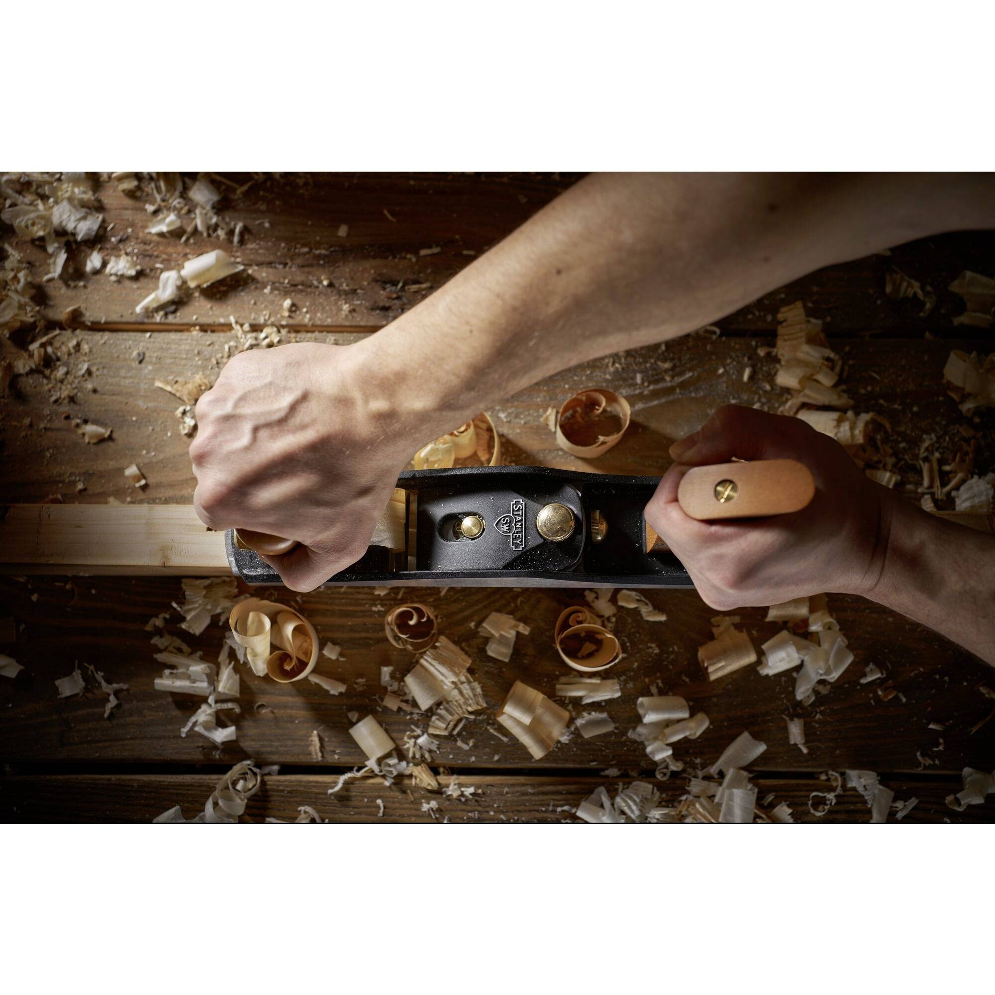 Two hands are using a plane to smooth wood on a table. Wood shavings are scattered, indicating craftsmanship.