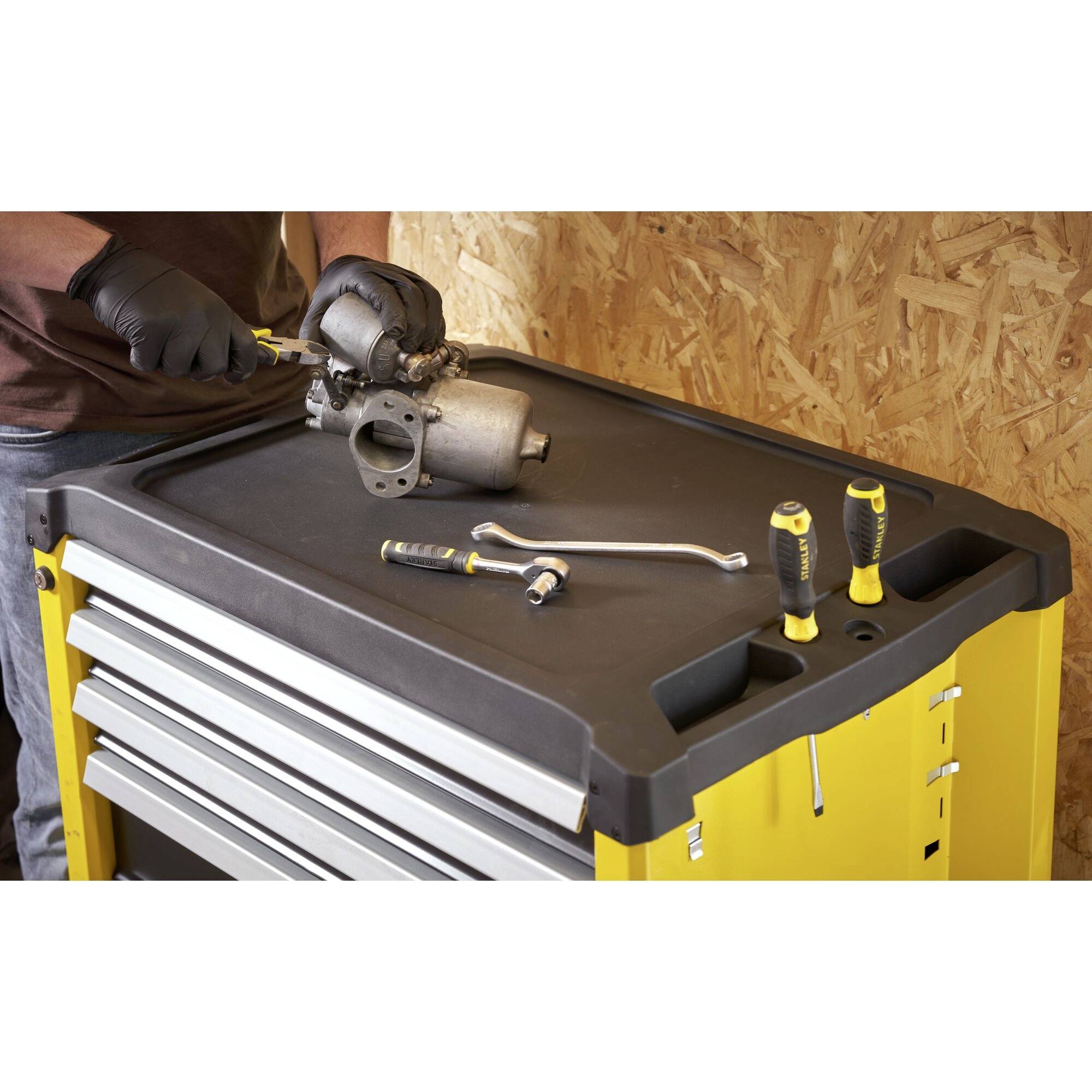 A person is repairing a car part on a yellow workshop trolley, surrounded by tools such as spanners and screwdrivers.