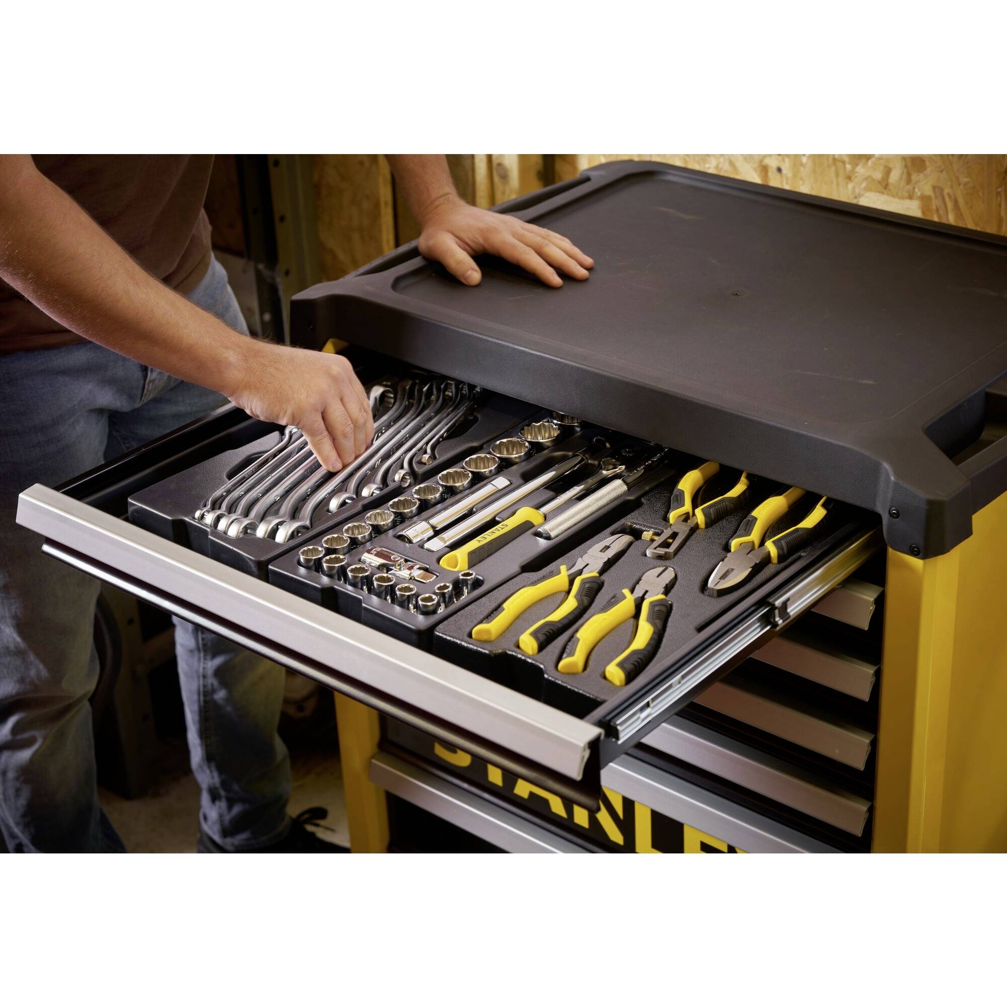 A person opens a drawer of a yellow tool trolley with various sorted tools such as spanners and pliers.