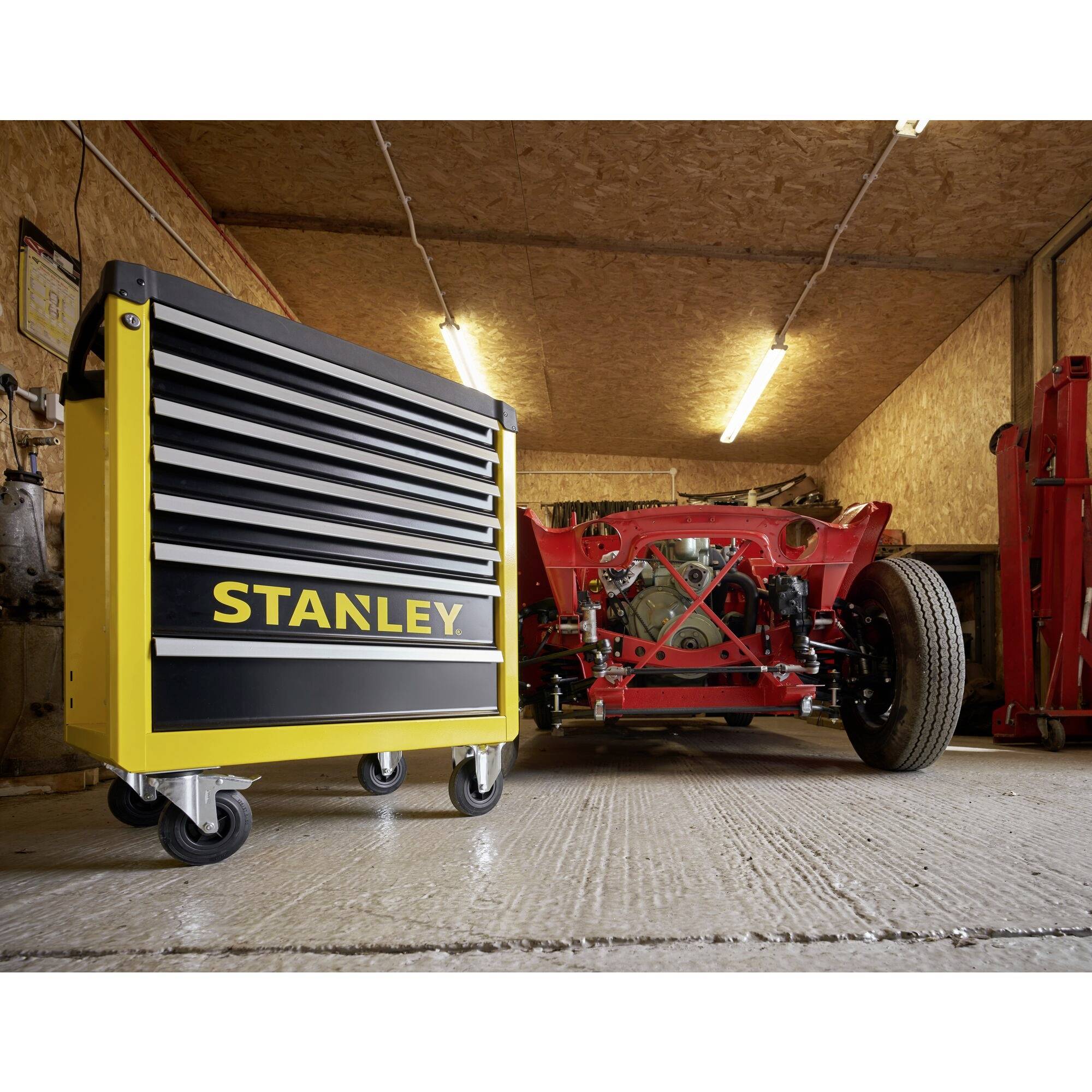 'Workshop with a yellow Stanley tool trolley on wheels in the foreground, red vehicle chassis in the background. Wooden panelling on the walls.'
