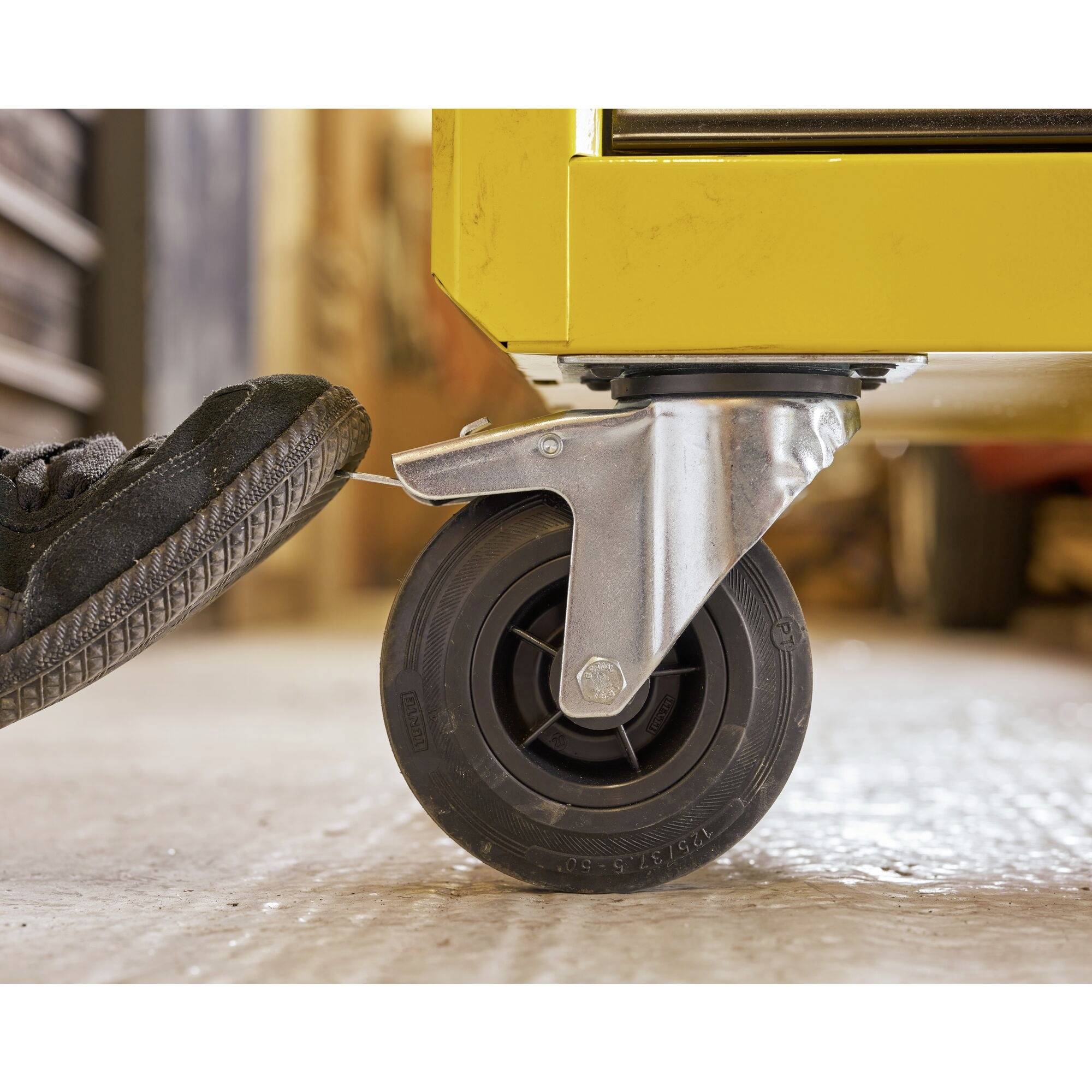 A foot presses on the wheel of a yellow vehicle standing on a concrete floor, in order to activate the brake.