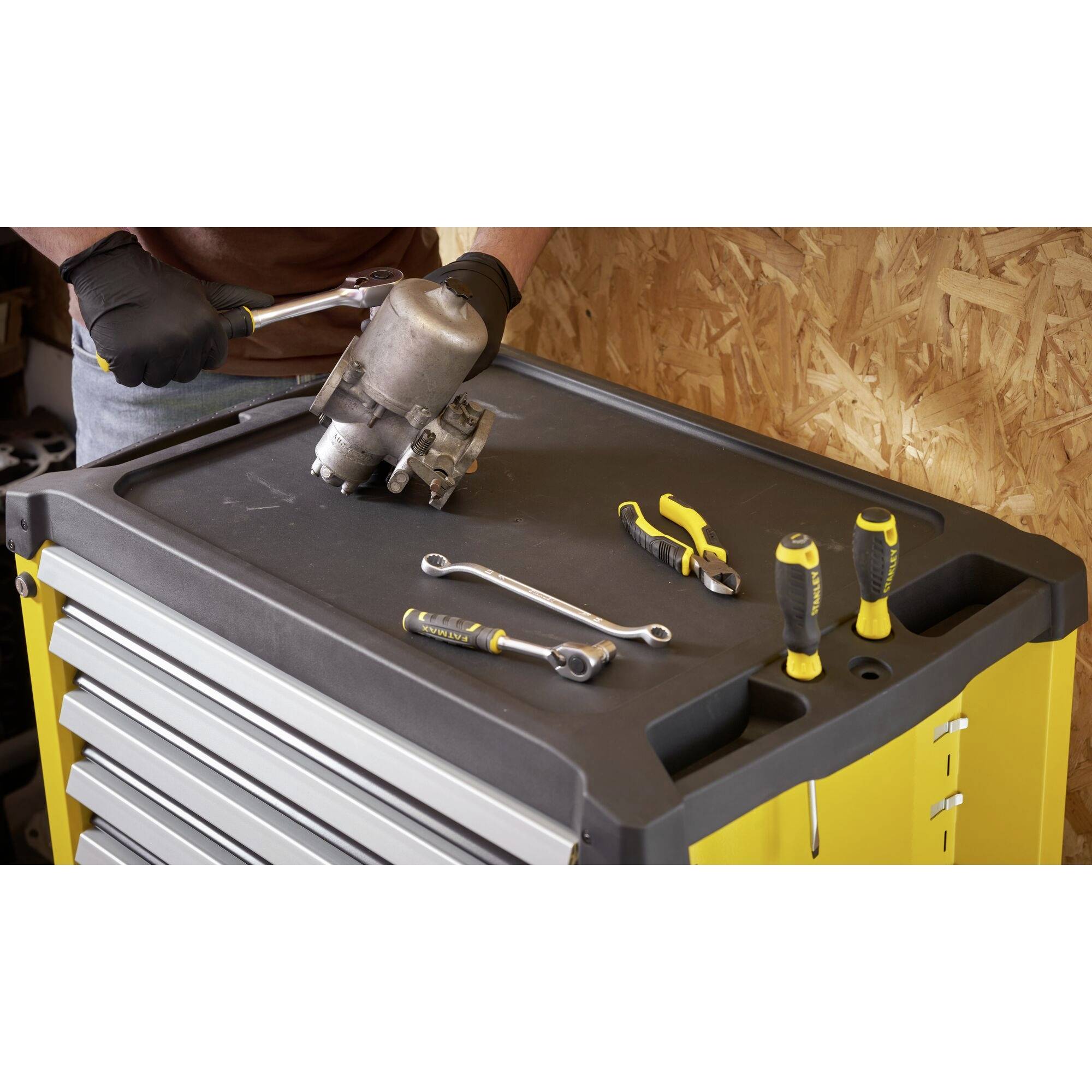 A person is repairing a metal part on a yellow toolbox. Various tools are laid out, including spanners and pliers.