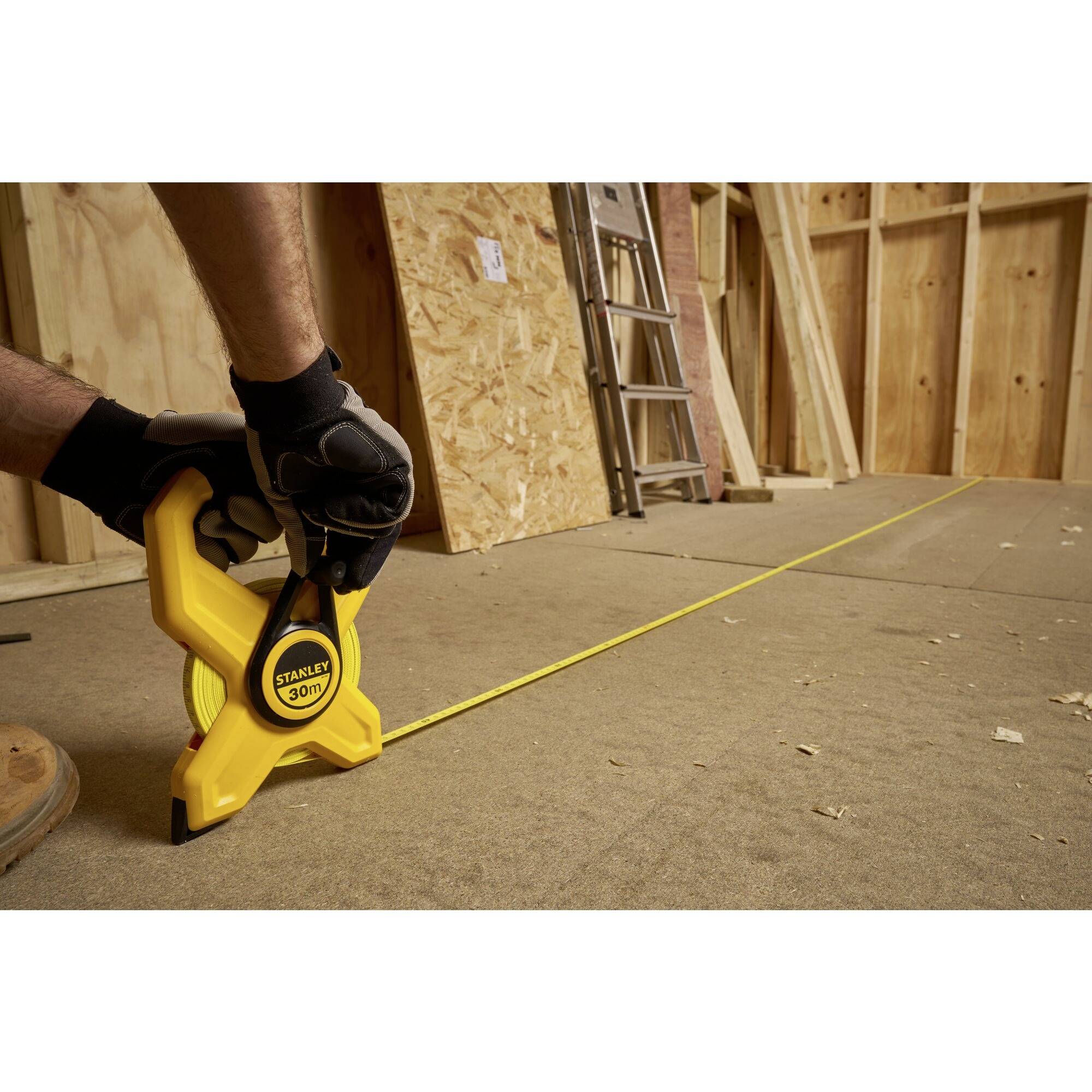 A person is measuring the distance on the floor in an unfinished building using a yellow measuring tape. A ladder is standing in the background.