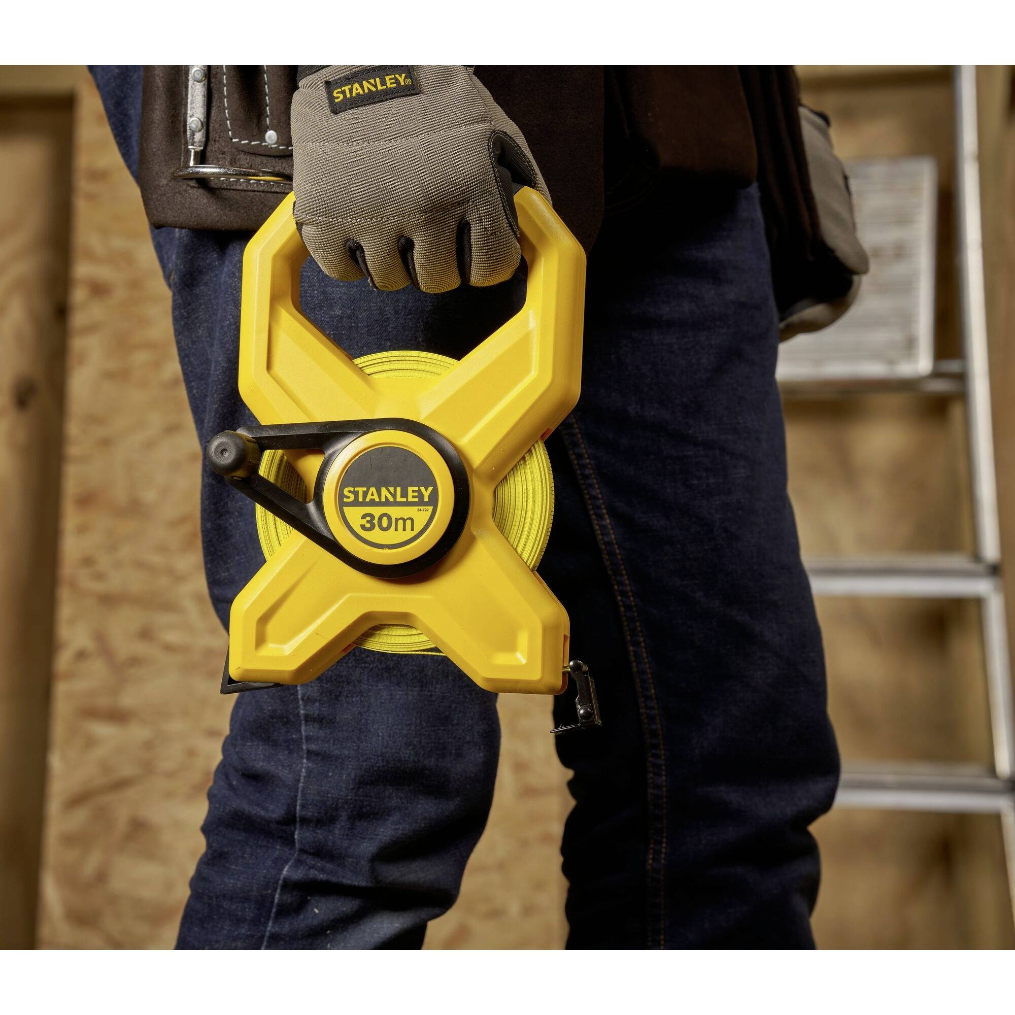 A person is holding a yellow tape measure with the inscription 'Stanley 30m' in their hand. A ladder is visible in the background against a wooden wall.