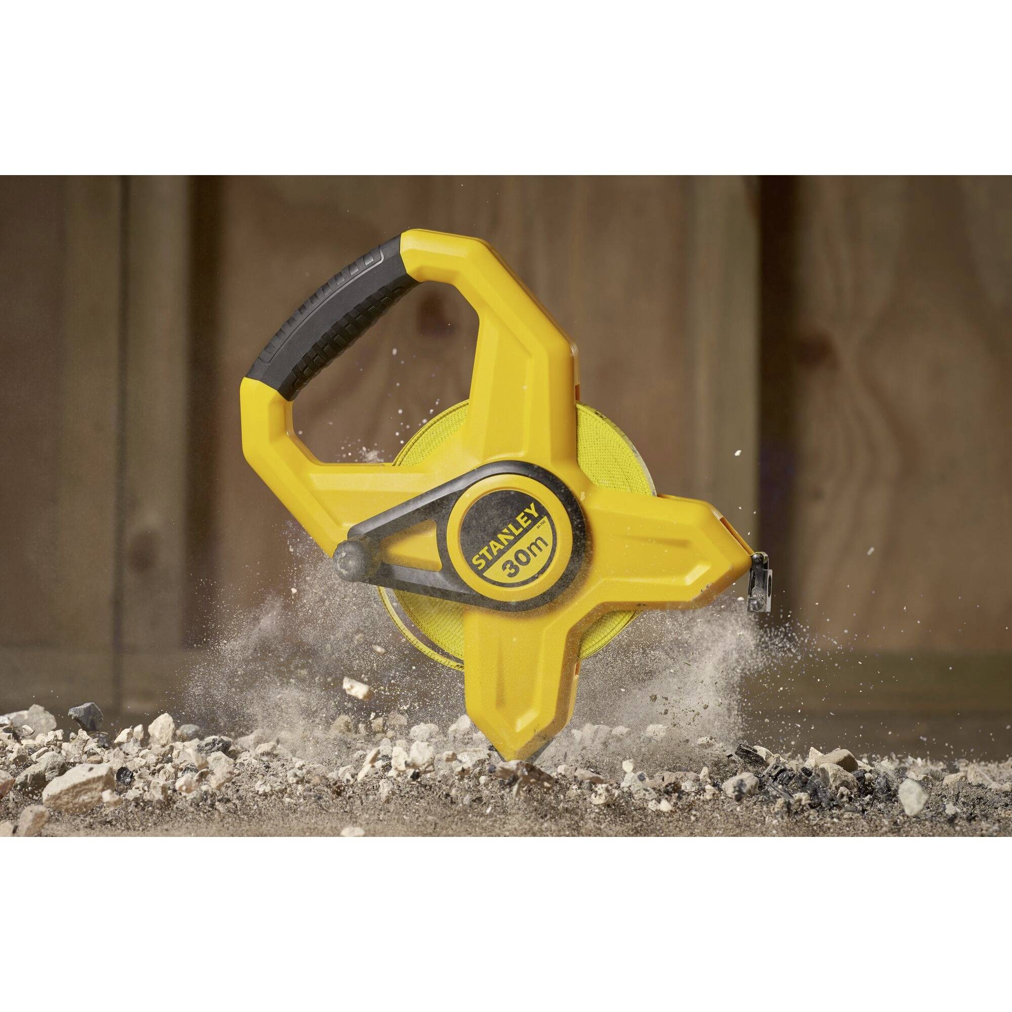 Yellow tape measure with a black handle, brand 'Stanley', lies on a concrete floor with dirt, with a wooden wall in the background.