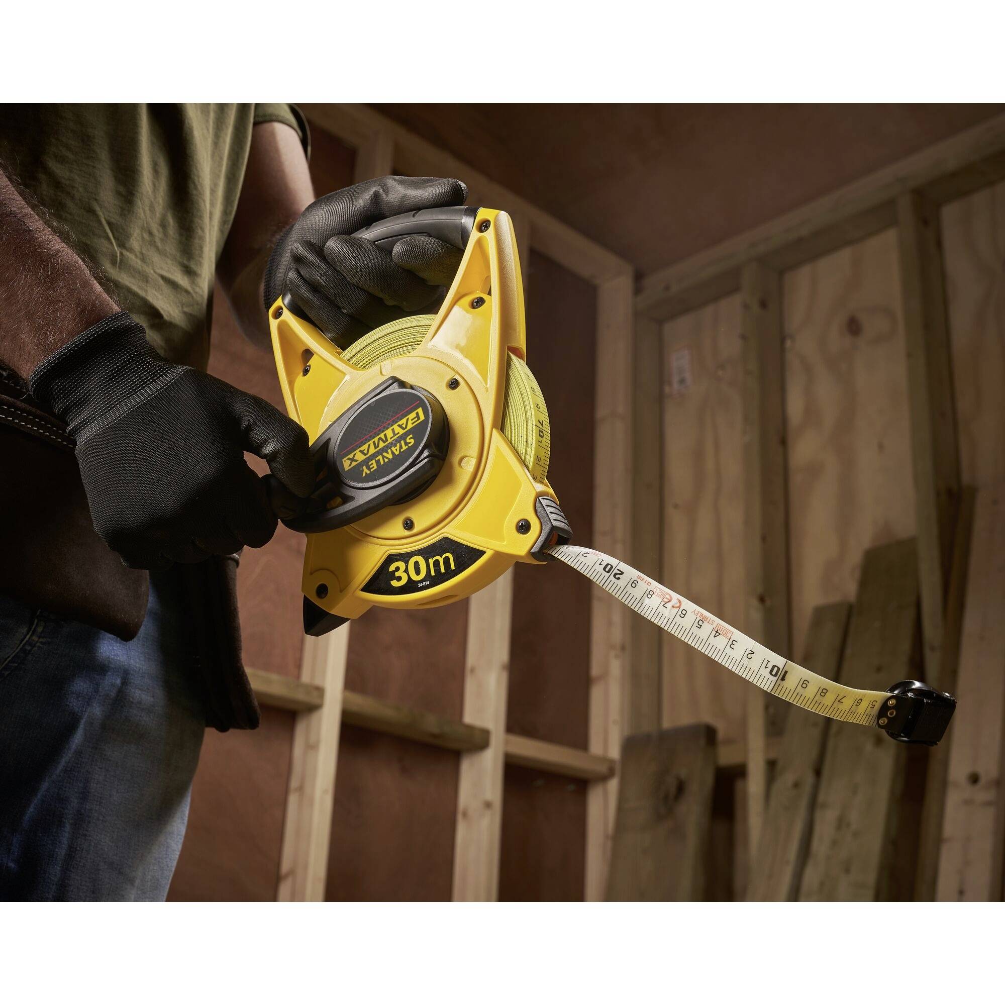 A man holds a yellow measuring tape with a 30-metre length in a wooden construction. He is wearing gloves, ready to take measurements.