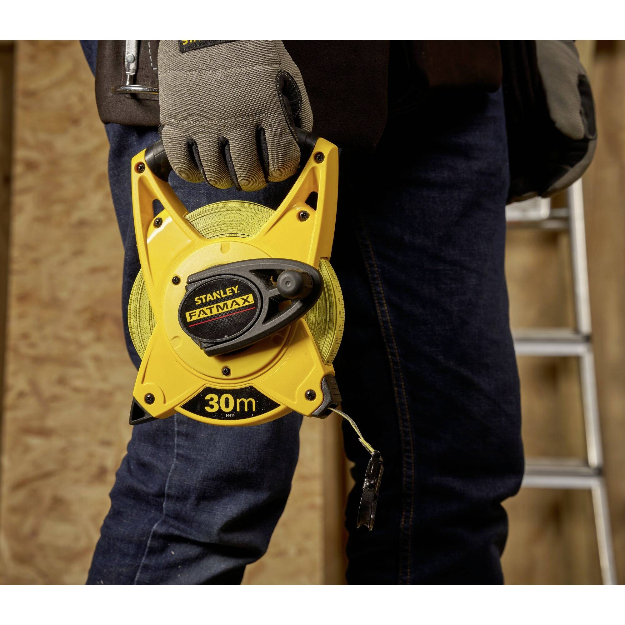 A person is holding a yellow 30-metre Stanley measuring tape in their hand. A ladder and wooden walls are visible in the background.
