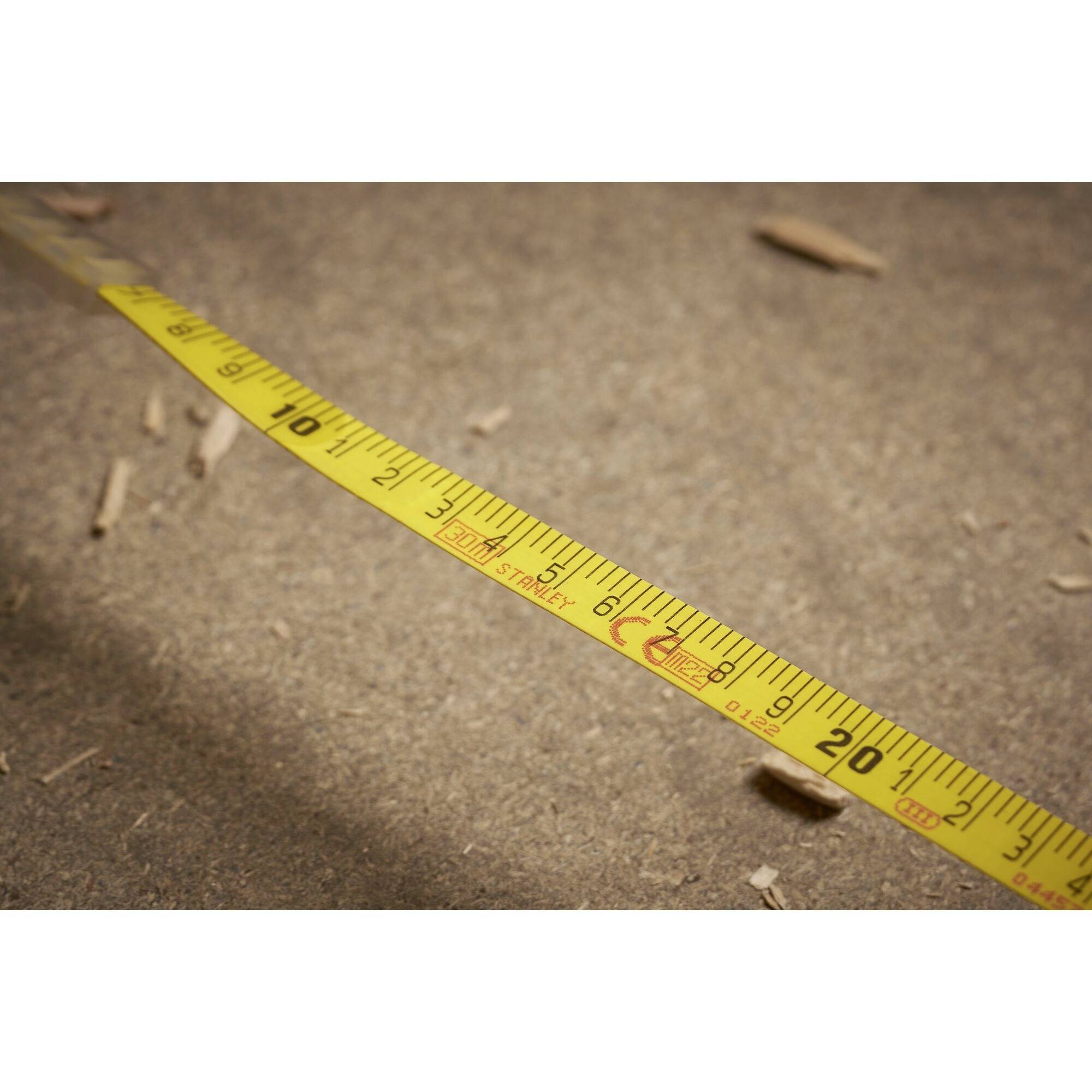Yellow tape measure, diagonally positioned on sandy ground. Measurements from 0 to 20 inches visible. Background shows wood shavings.