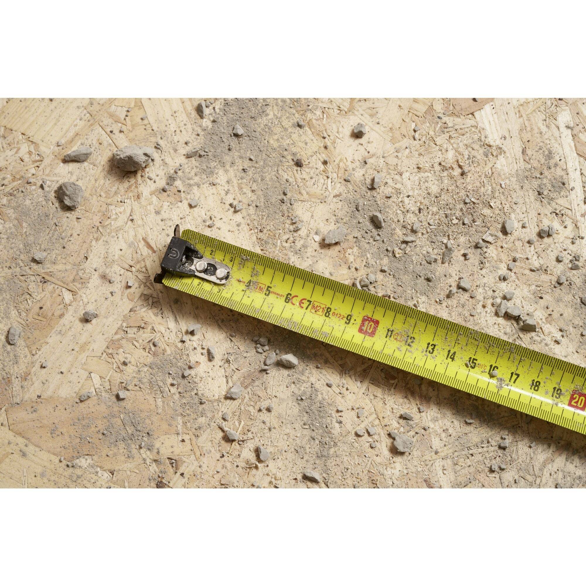 A yellow tape measure lies on a dusty wooden floor, surrounded by small stones and wood splinters.