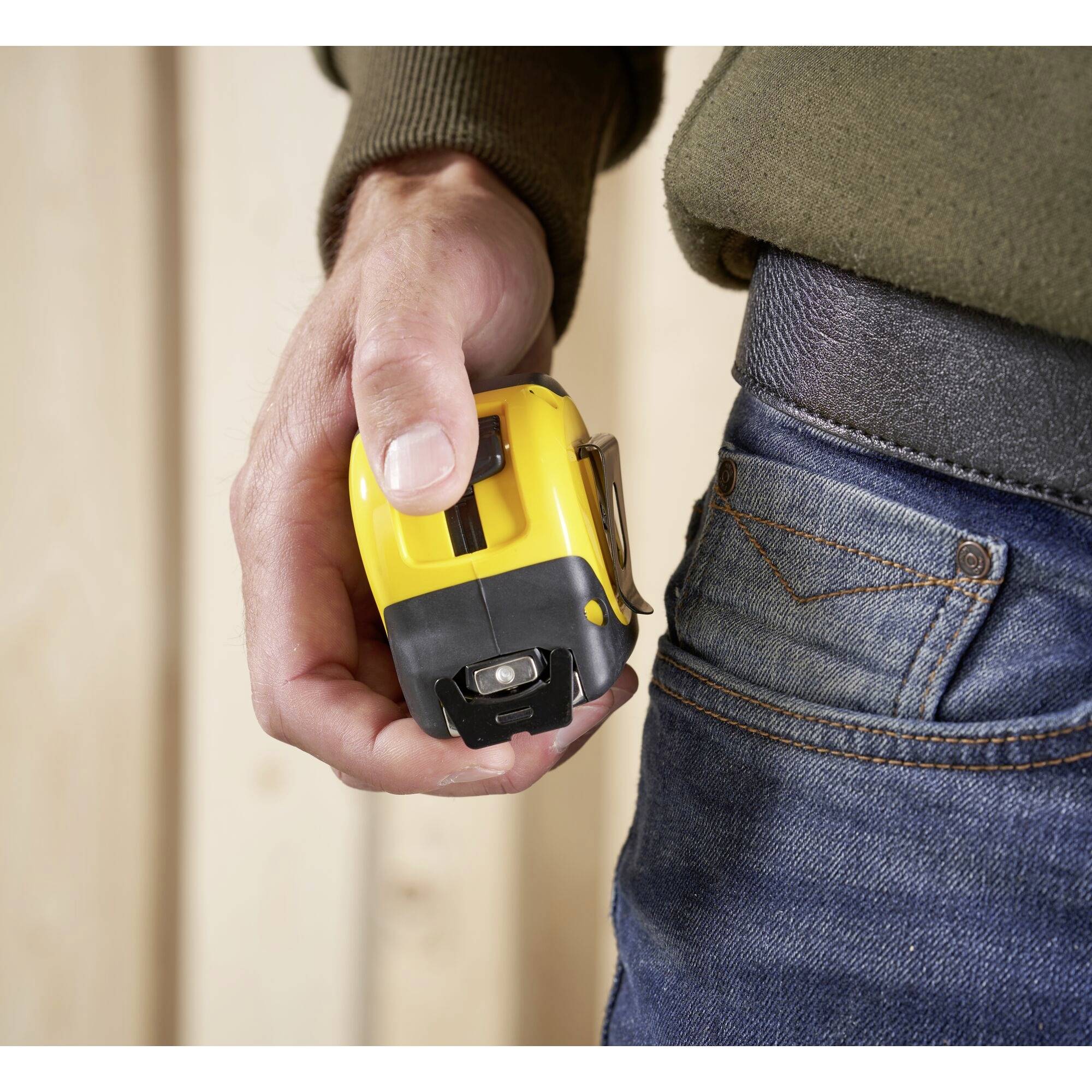 A person is holding a yellow tape measure near a wooden wall. The person is wearing blue jeans and an olive green jumper.