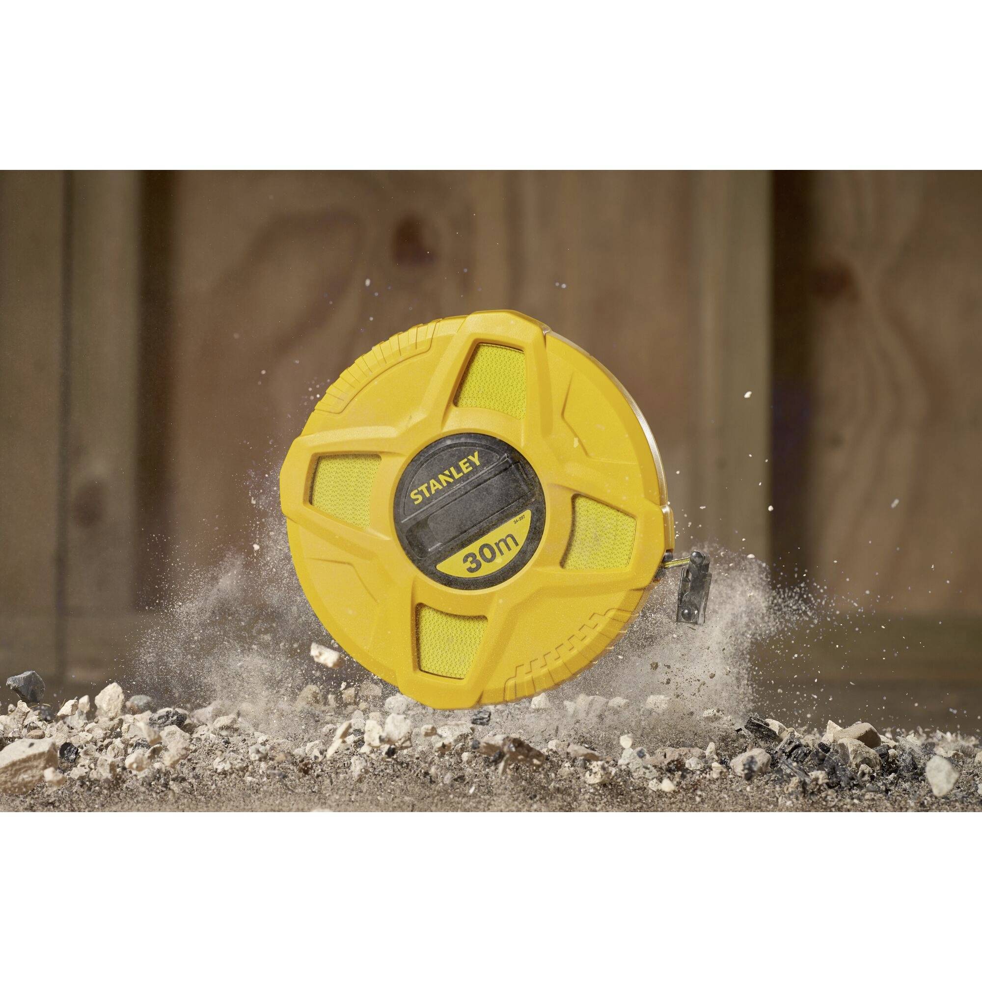 A yellow tape measure with '30 m' printed on it lies on gravel and sand. Wooden background. Brand name 'Stanley' on the tape measure.