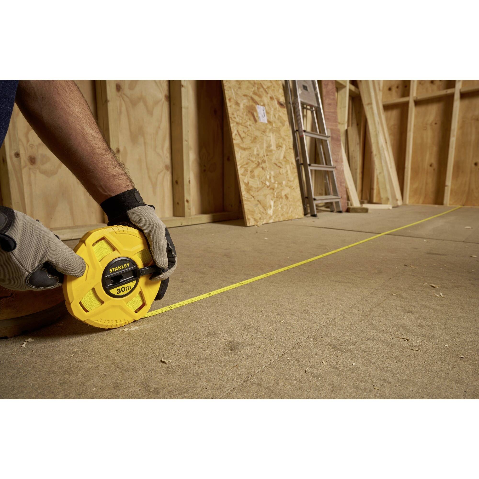 A person is measuring the floor with a yellow tape measure in an unfinished room with wooden walls. A ladder is standing in the background.