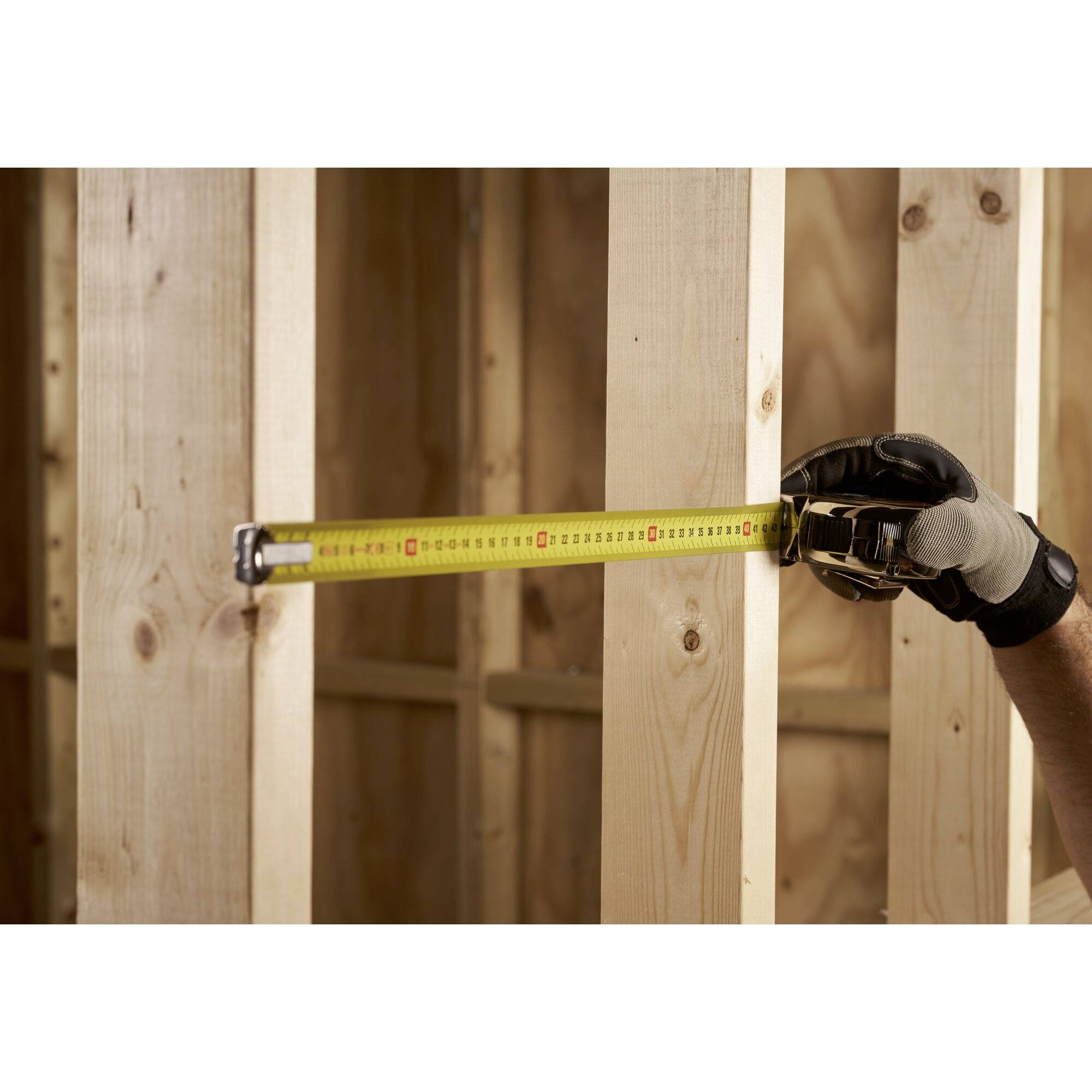 'A person wearing a glove is holding a tape measure against a wooden structure, measuring the distance between two vertical wooden slats.'